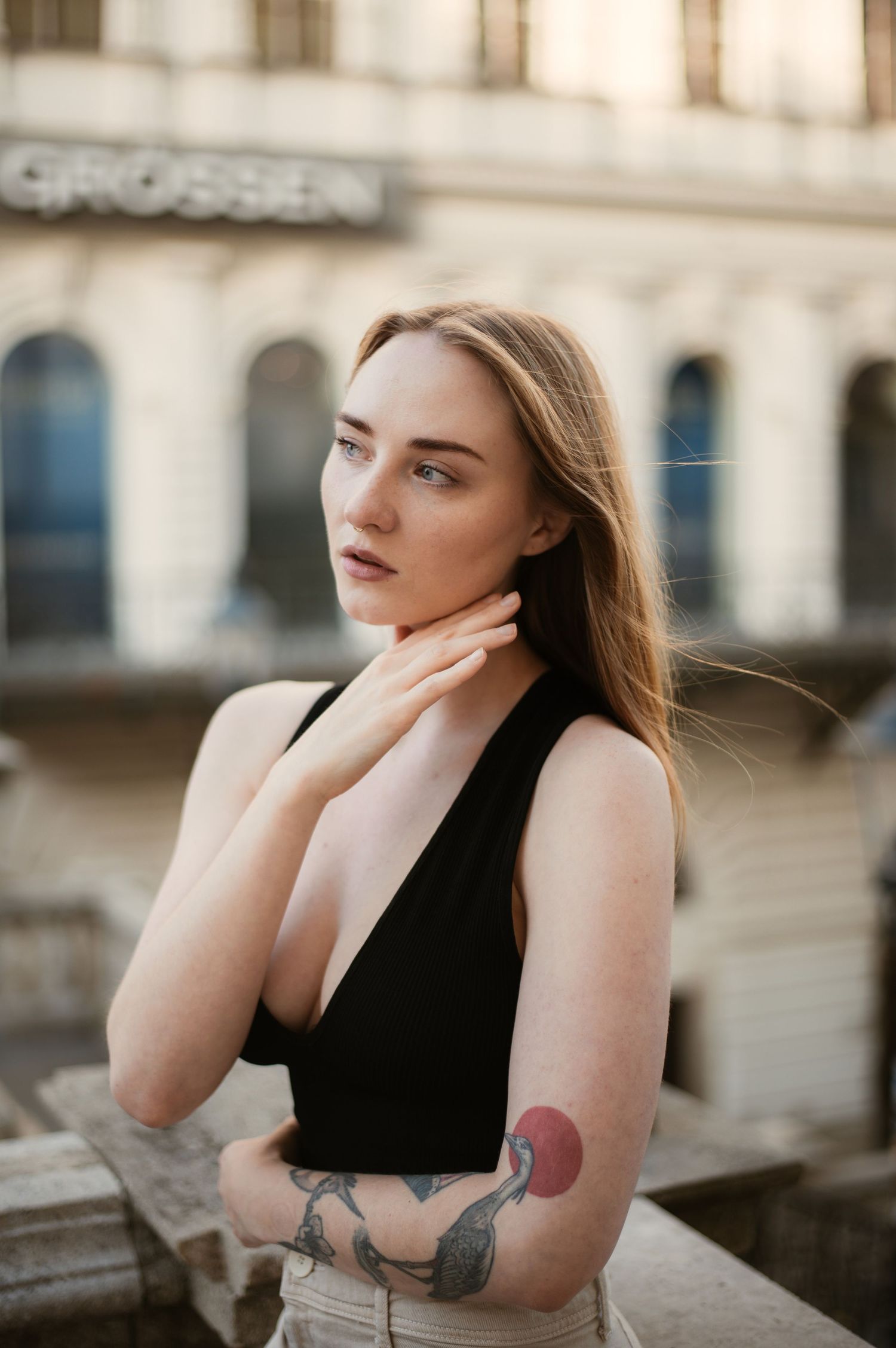 Blonde woman with arm tattoo in black sleeveless top poses by stone railing near European building.