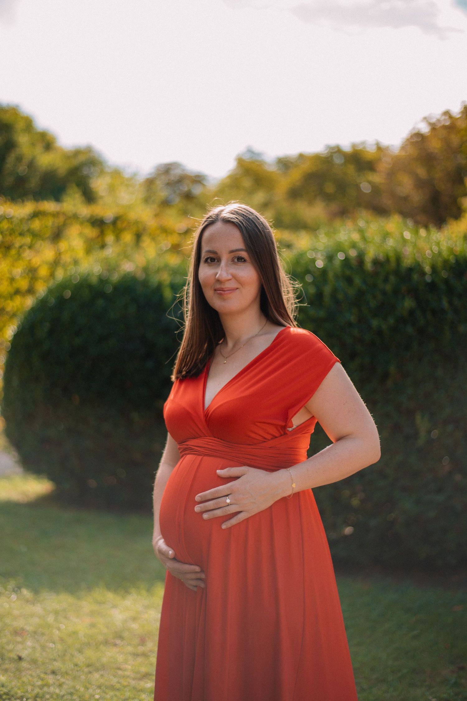 Pregnant woman in red dress cradling baby bump outdoors in lush green garden during golden hour.