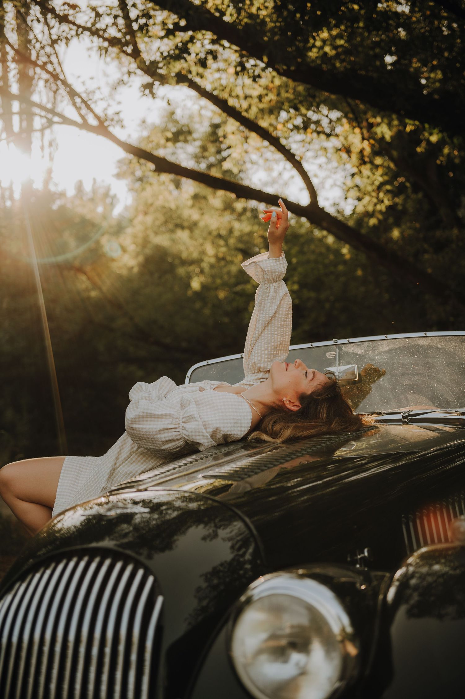 Woman in white dress lying on vintage classic car hood at golden hour sunset with trees in background.