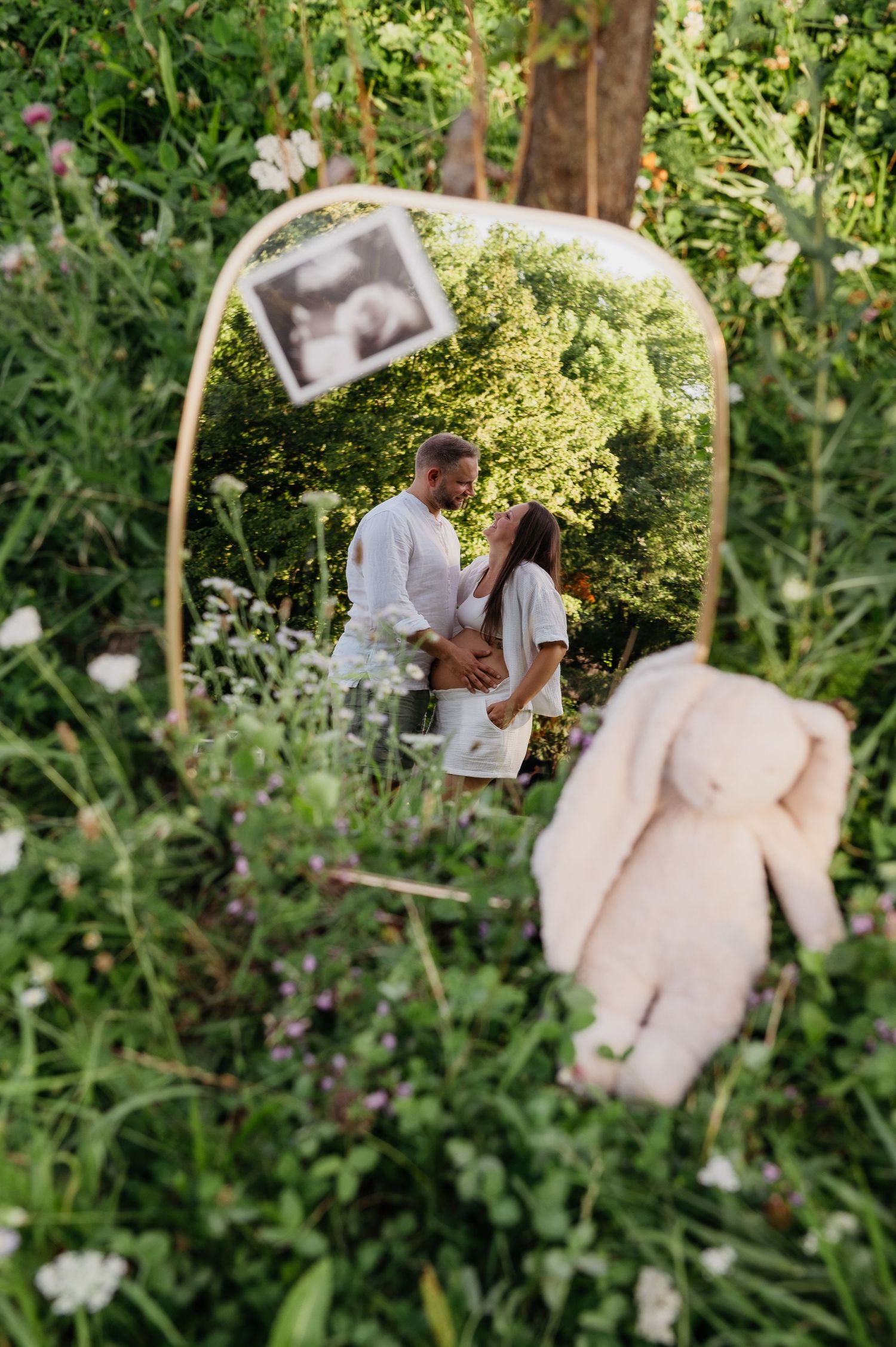 A couple embraces in a garden, reflected in an oval mirror surrounded by wildflowers, with a plush bunny and ultrasound photo nearby.
