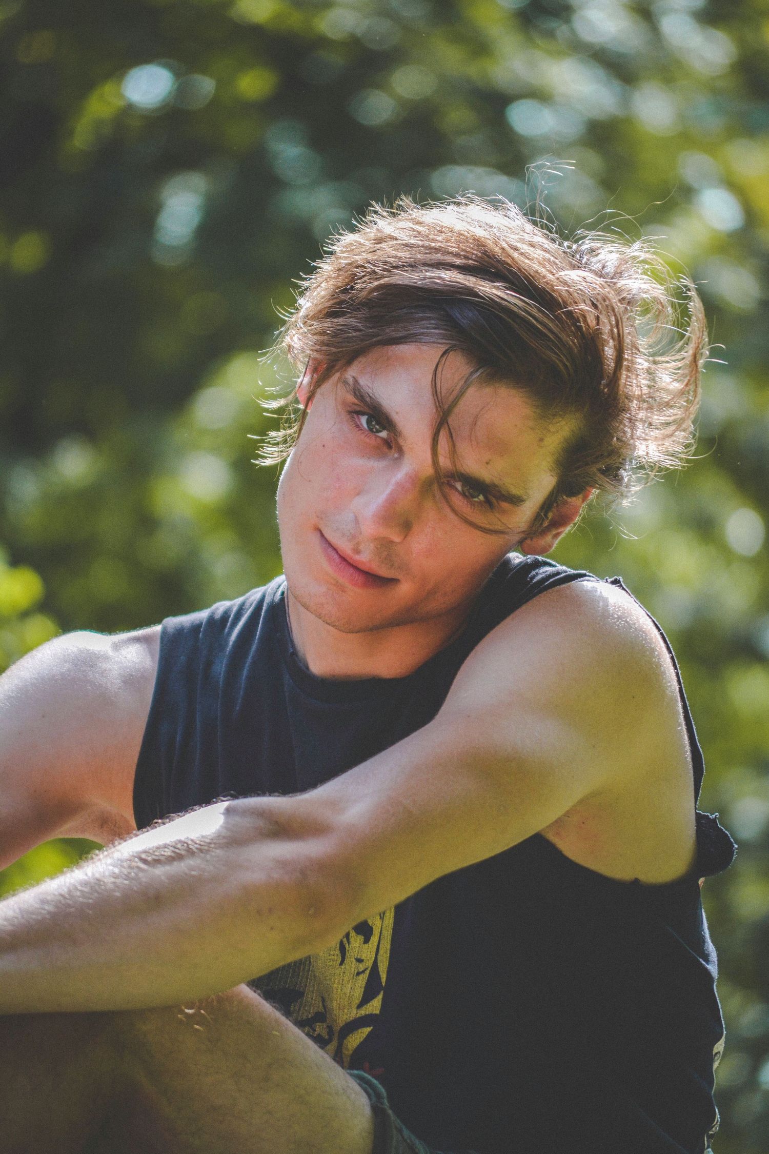 Young man with brown hair in black sleeveless shirt sitting outdoors with arms crossed, looking intensely at camera.