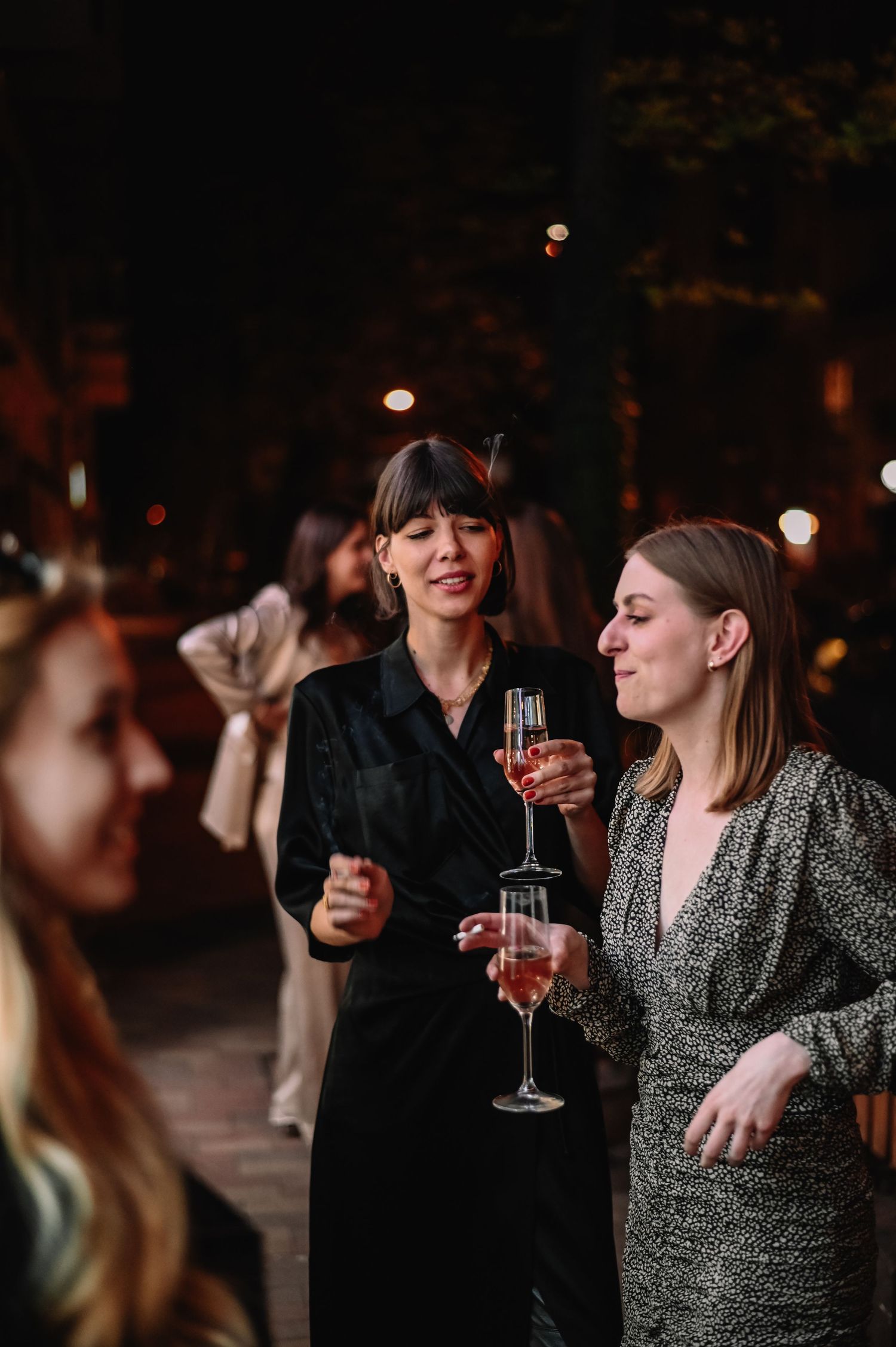 Two women in elegant dresses holding champagne glasses at an outdoor evening party.