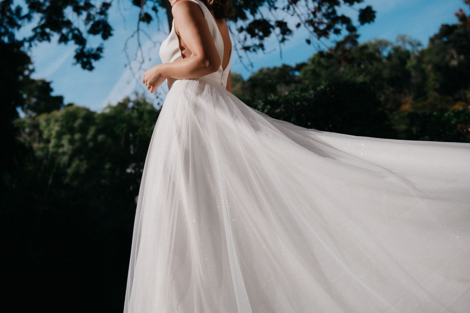 Bride in flowing white tulle wedding gown twirling outdoors with trees and blue sky in background.