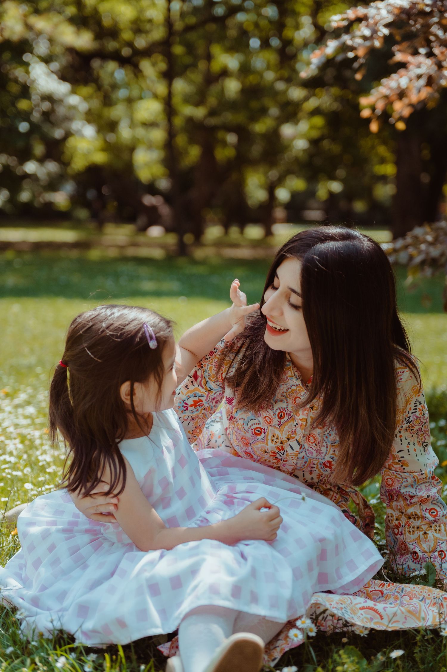A mother and young daughter share a tender moment sitting on grass in a sunny park, both wearing floral dresses.