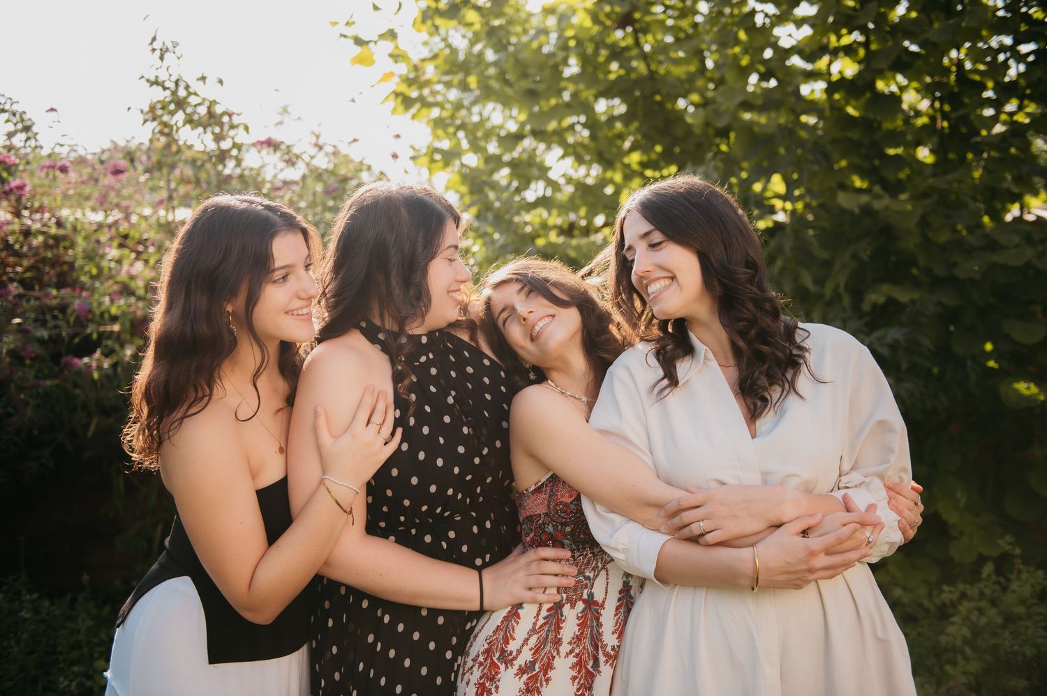 Four young women laughing and embracing outdoors in golden sunlight, surrounded by lush green trees.