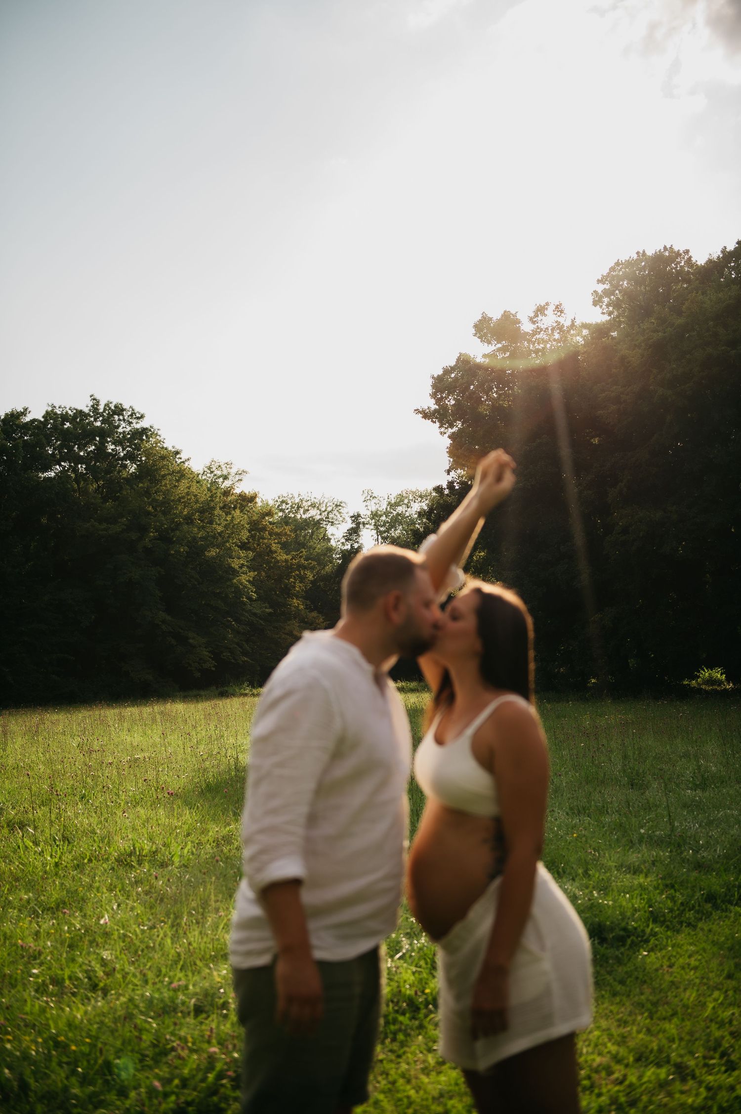 Pregnant couple sharing a tender kiss outdoors in a sunlit green meadow surrounded by trees during golden hour.