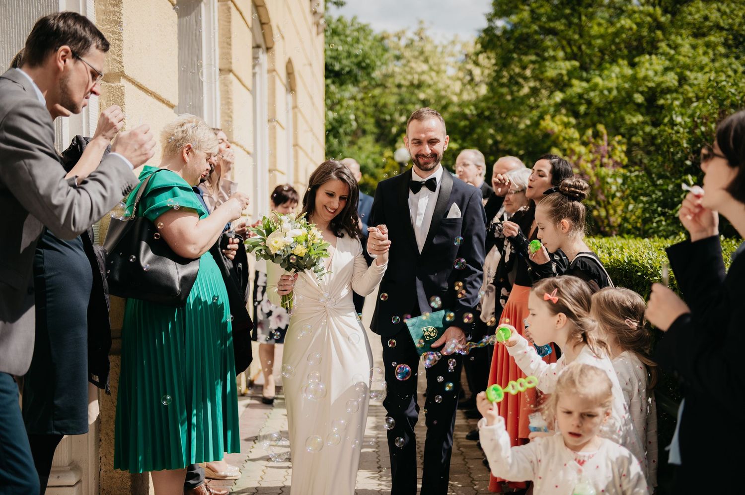 Newlyweds walk through joyful guests blowing bubbles outside a stone building on their wedding day.