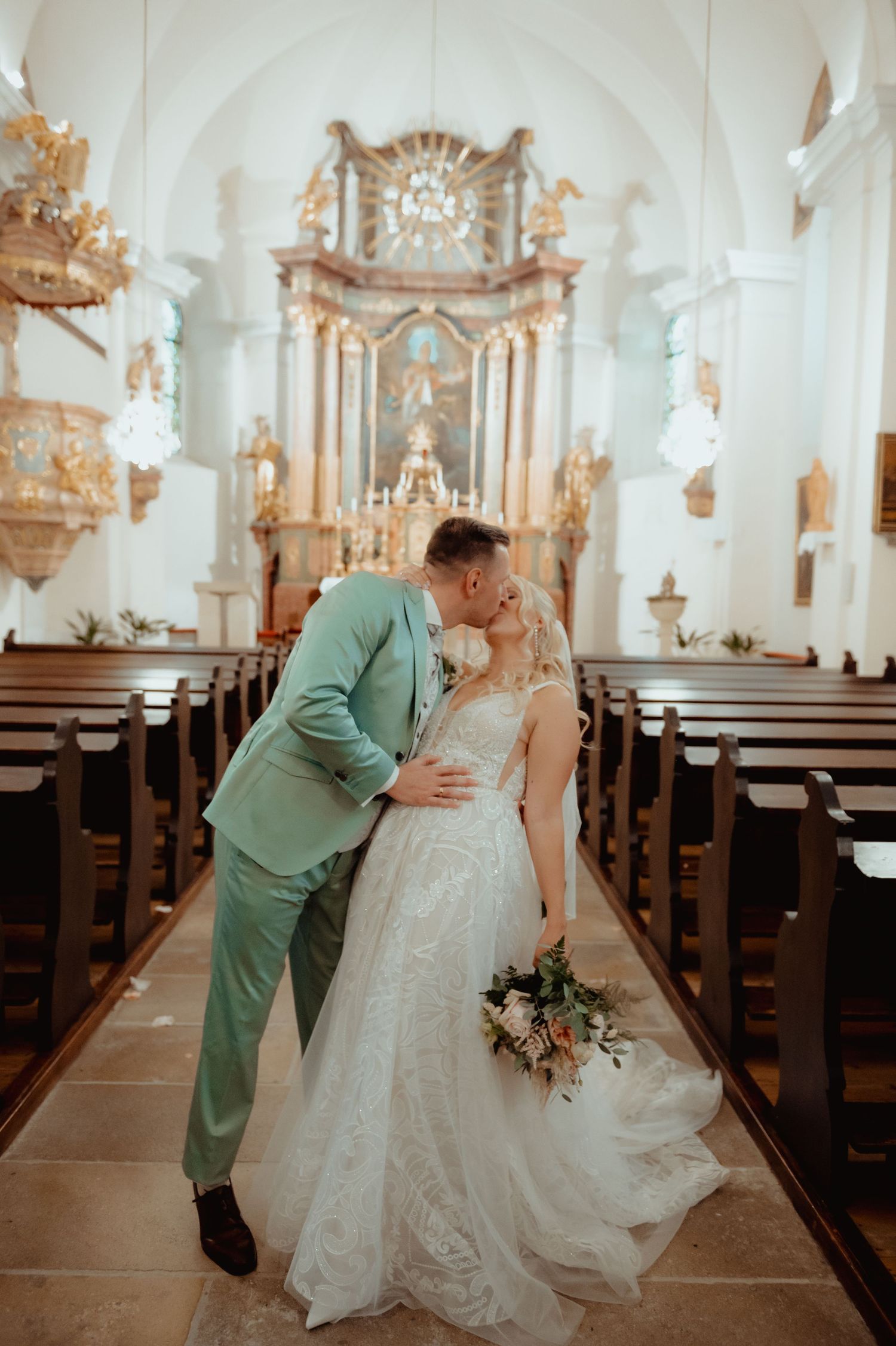 Bride and groom share a romantic kiss in an ornate white church aisle, bride holding a bouquet in a lace gown.
