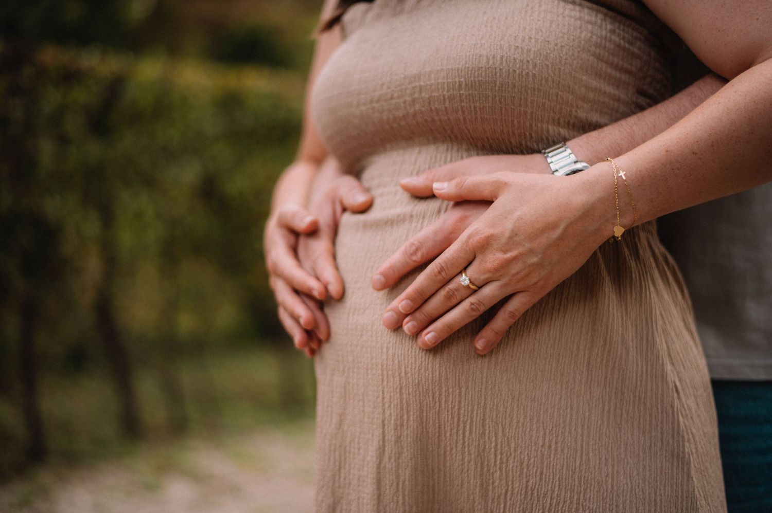 Close-up of a pregnant woman in a beige dress with hands tenderly cradling her baby bump outdoors.