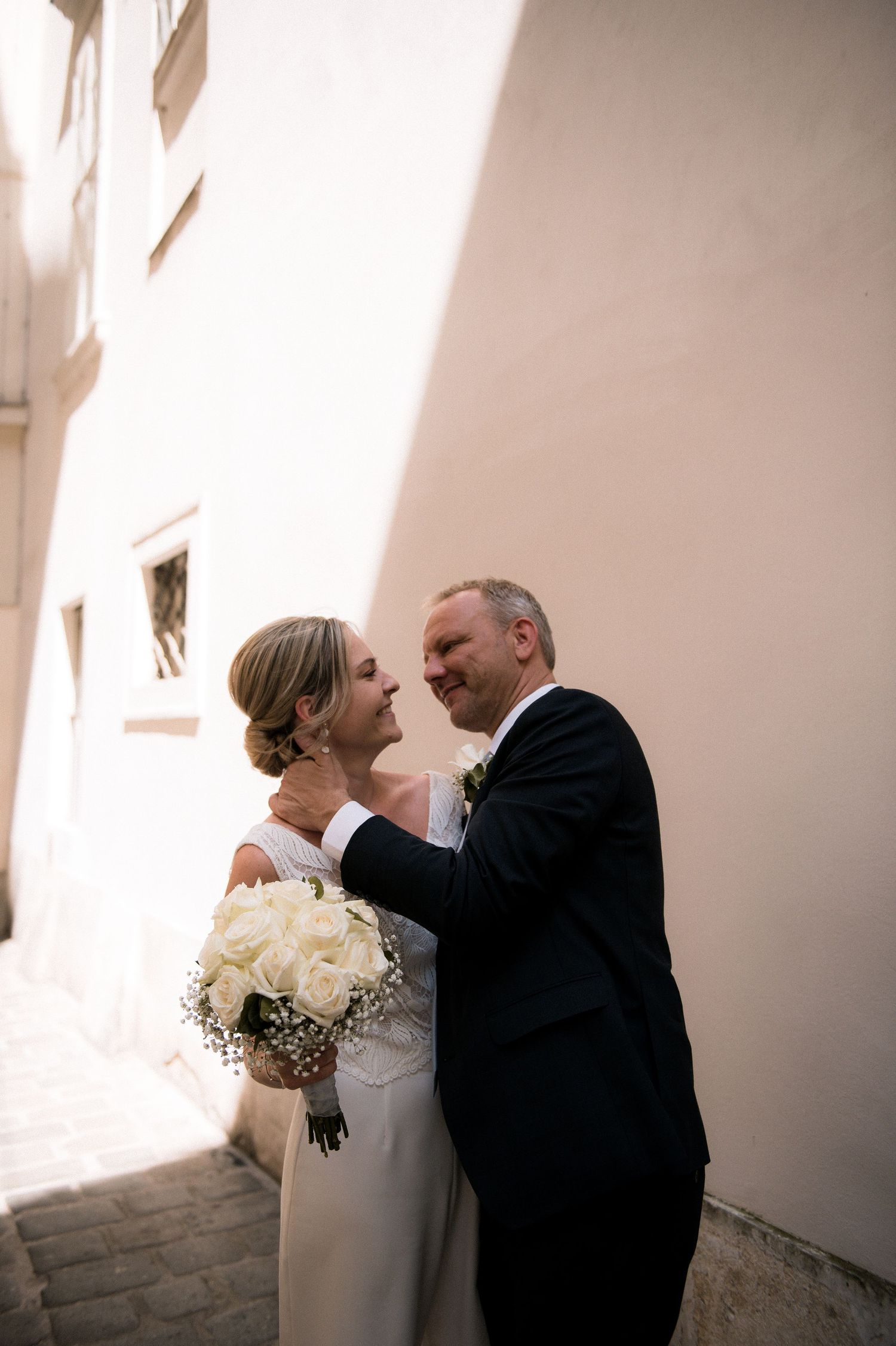 Bride and groom share a tender moment in a sunlit alley, bride holding white rose bouquet in elegant wedding attire.