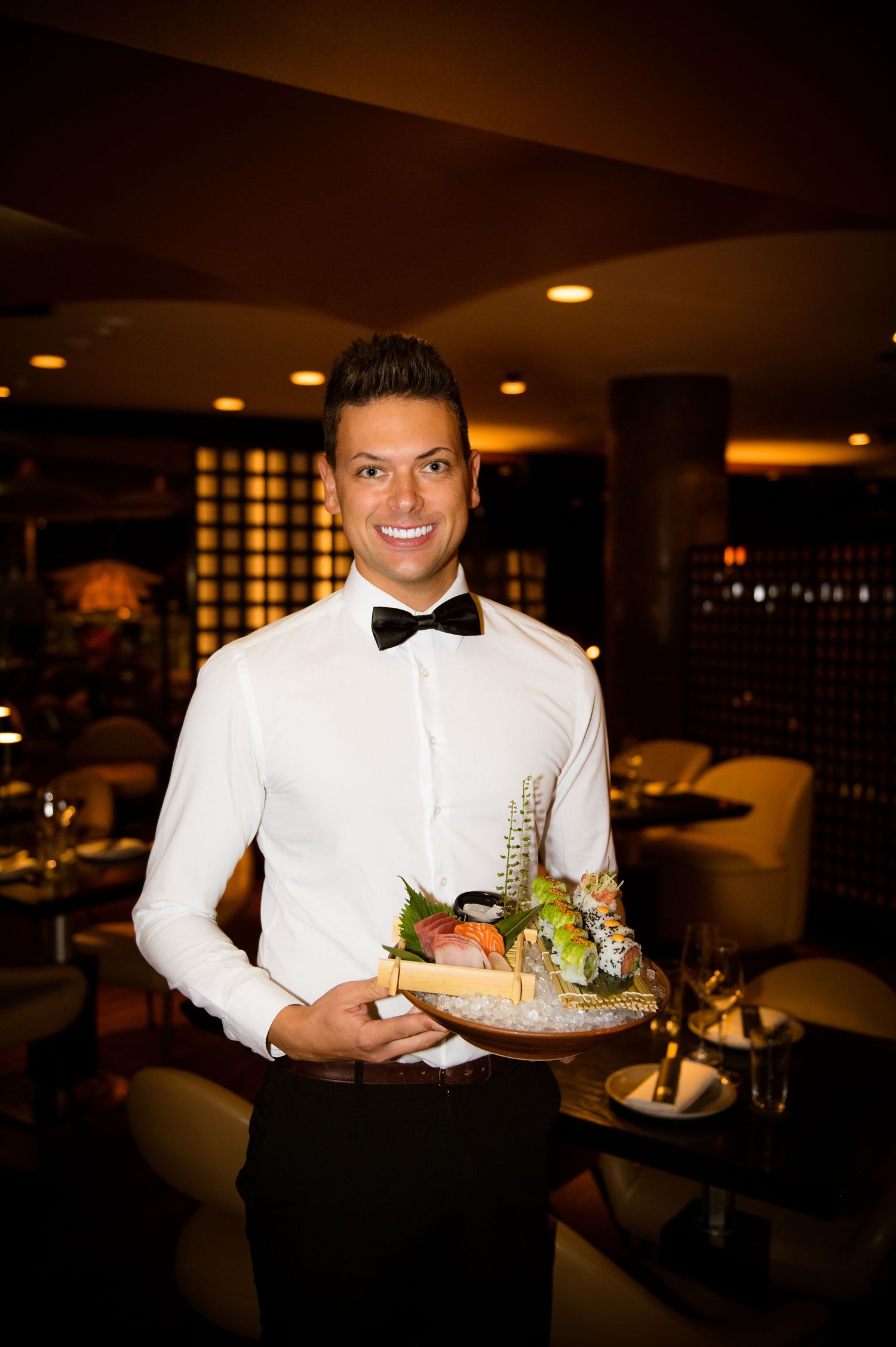 Smiling waiter in white shirt and bow tie holding a tray of gourmet food in an elegant restaurant.