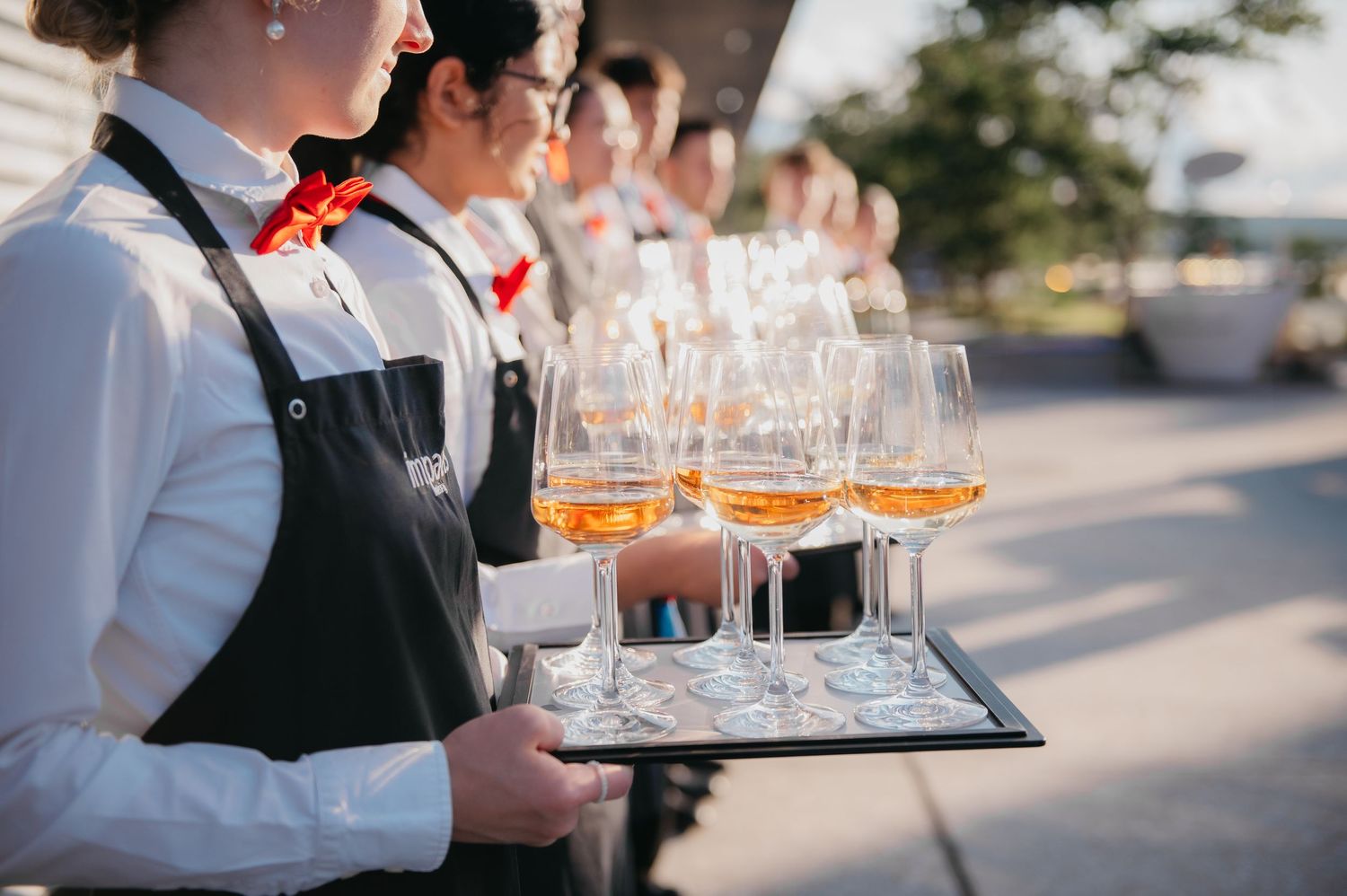 Waitstaff in black aprons serving amber cocktails in wine glasses on a silver tray at an outdoor event.