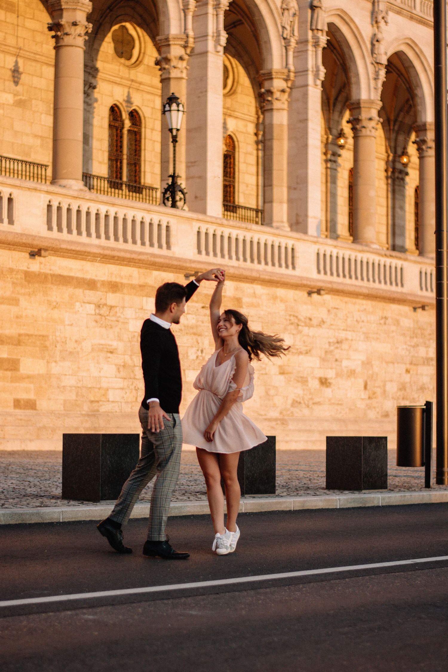 Couple dancing romantically on a cobblestone street at sunset in front of a grand historic building with arched columns.