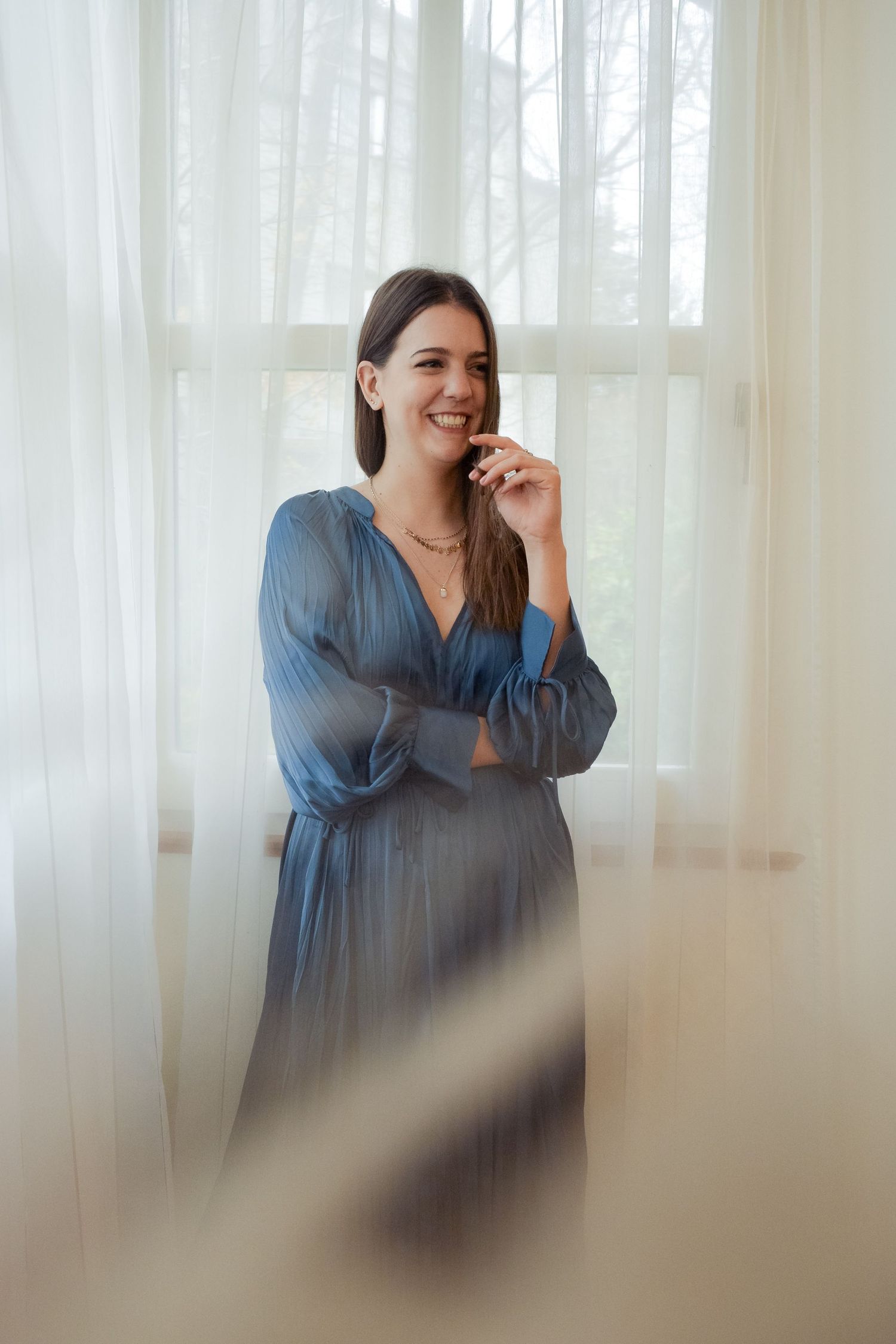 Smiling brunette woman in a blue satin dress stands by sheer white curtains in natural light.