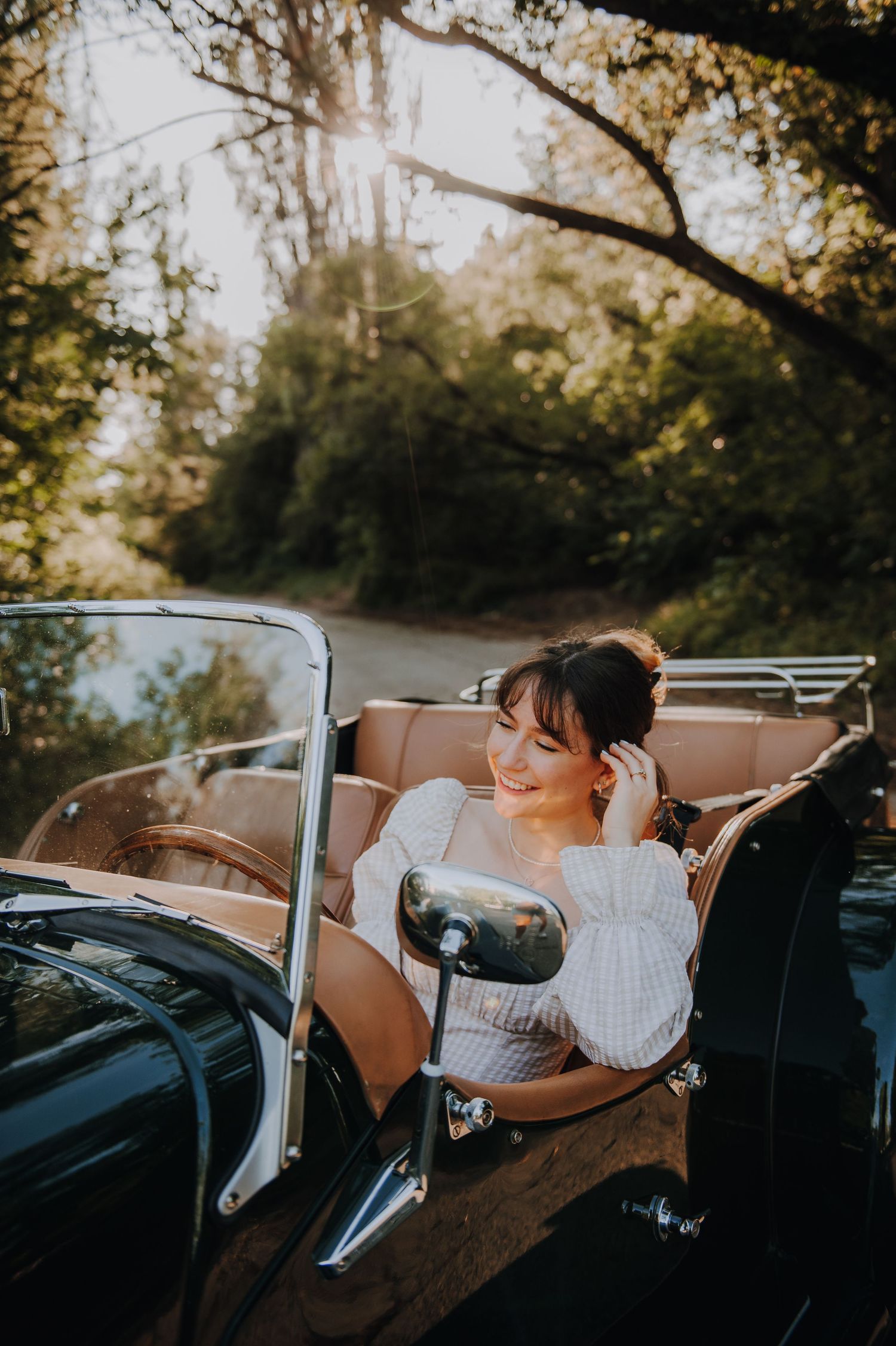 A smiling woman in a white dress sits in a vintage convertible car surrounded by lush green trees in golden sunlight.