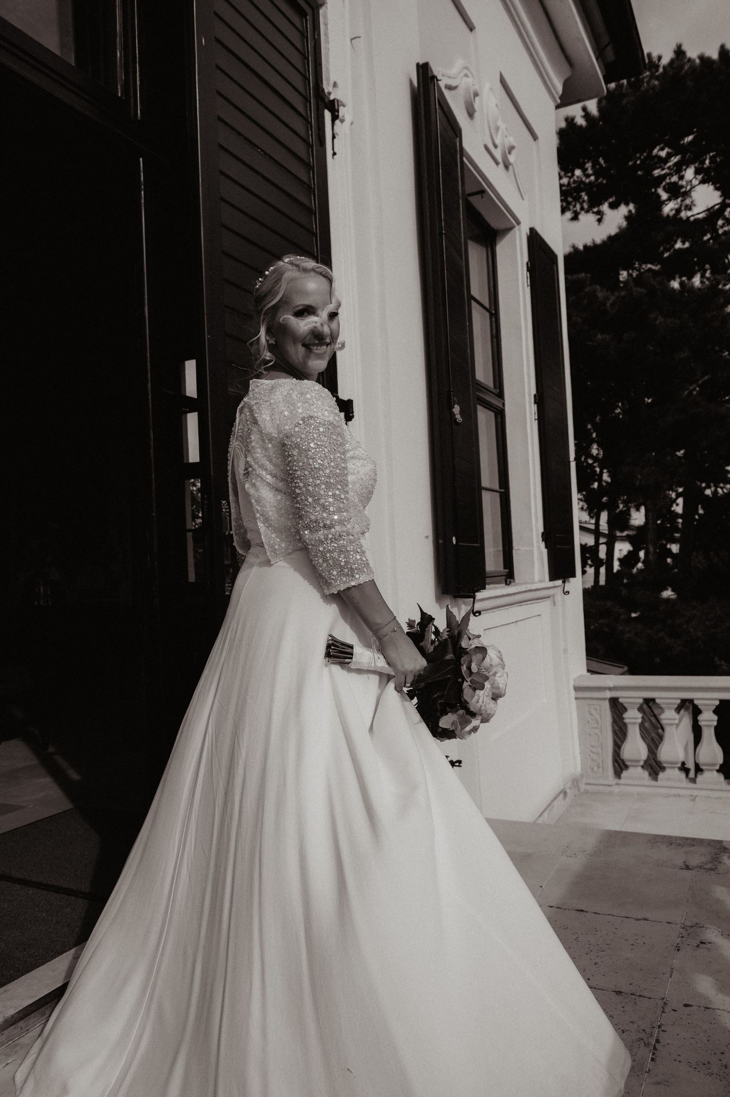 Bride in elegant pearl-embellished wedding gown holding bouquet, posing outside historic venue in black and white photo.