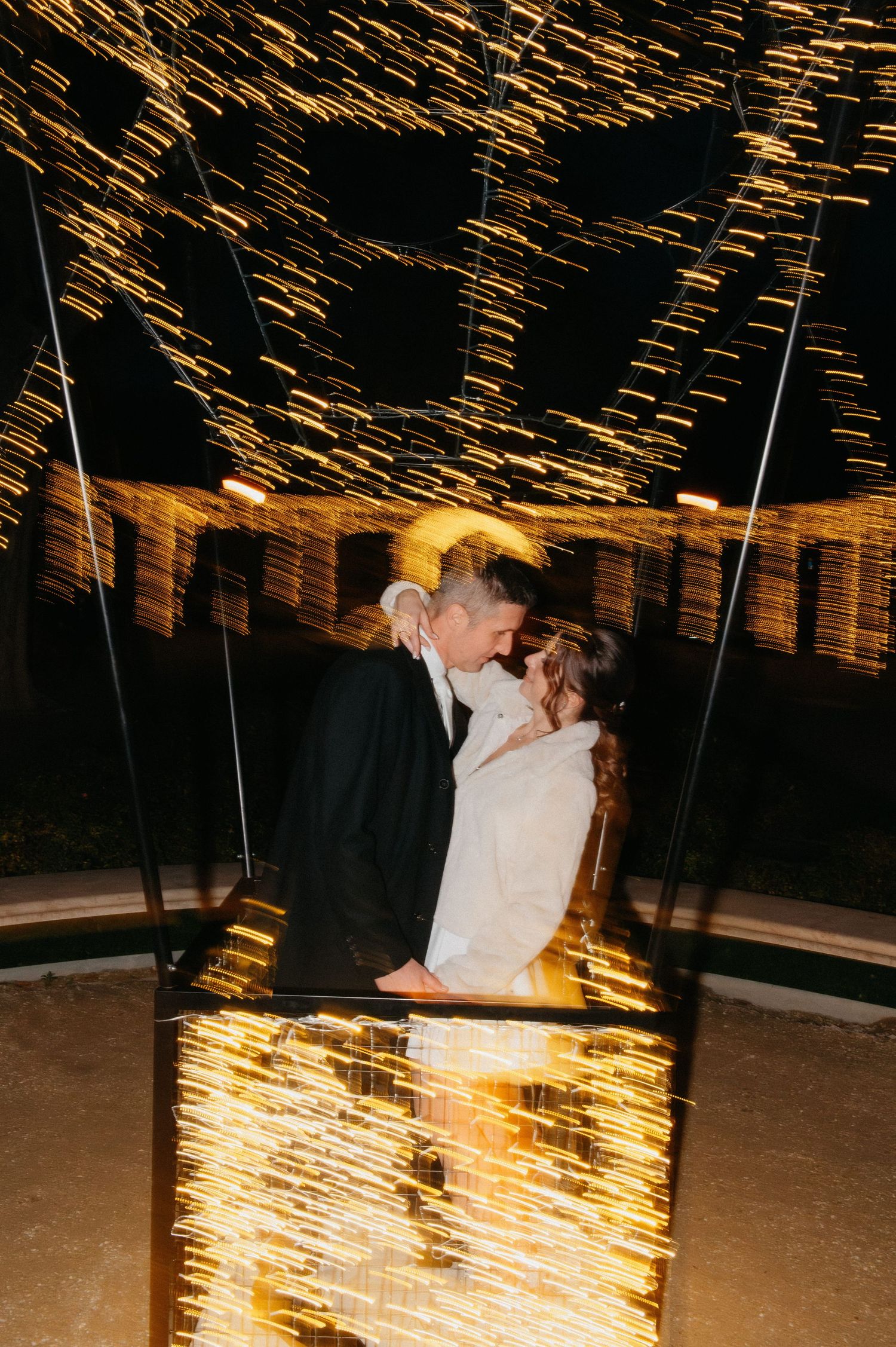 Bride and groom share a romantic kiss surrounded by stunning golden light streaks at night.