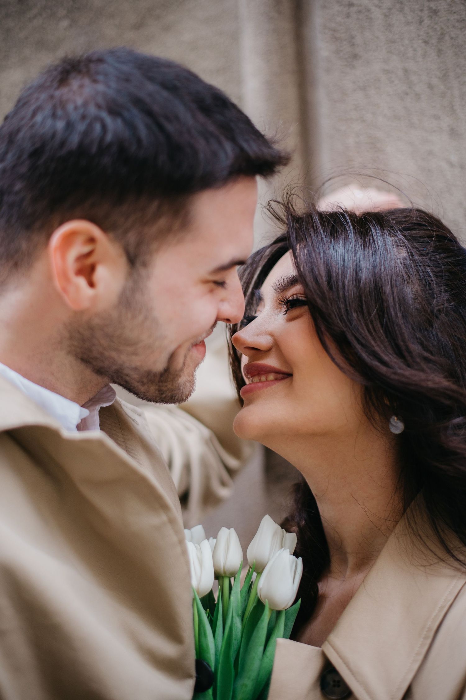 Close-up of a smiling couple nearly kissing, woman holding a bouquet of white tulips against a stone wall.