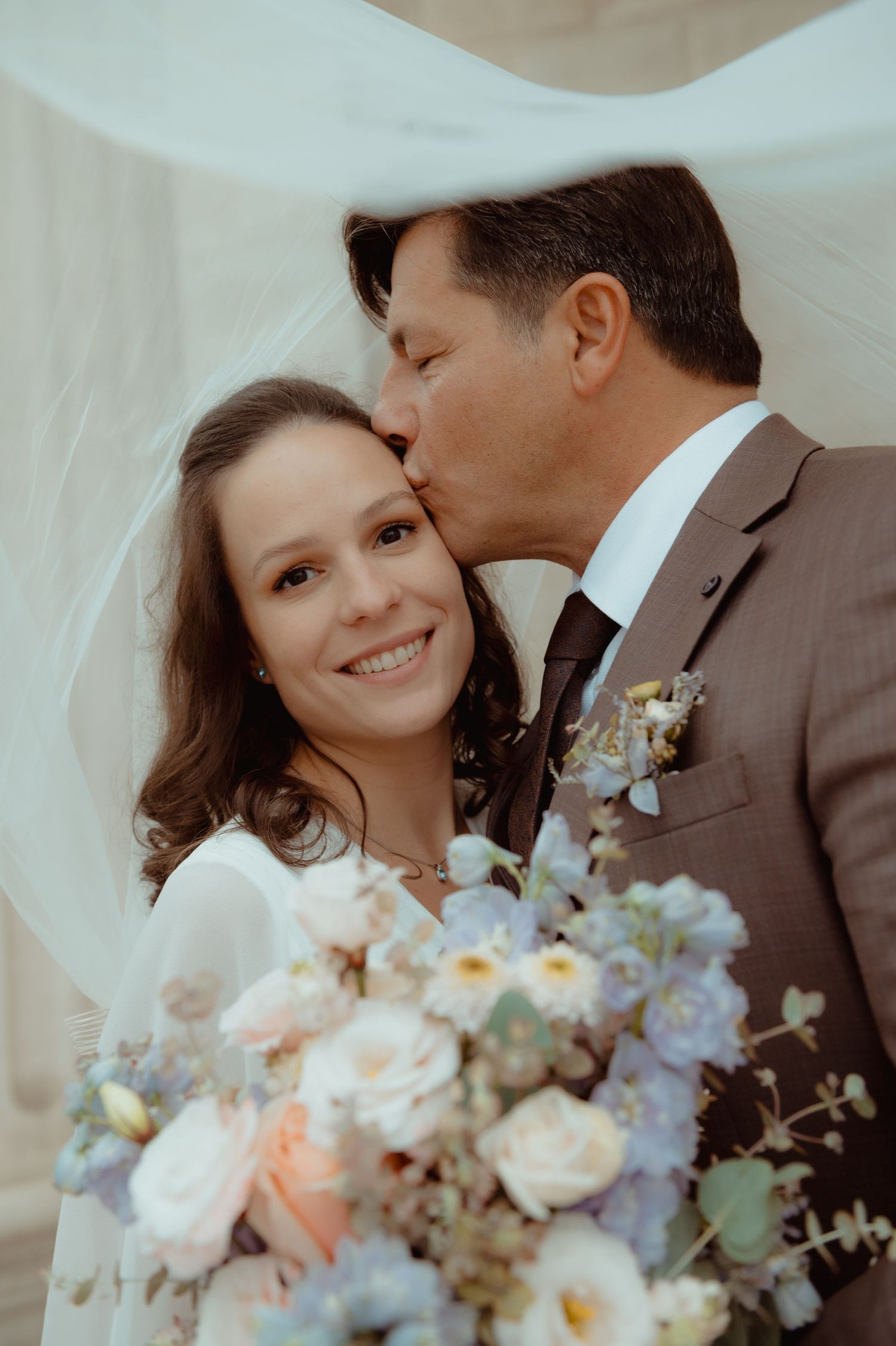 Groom in brown suit kisses bride on cheek as she smiles holding pastel floral bouquet under flowing veil.