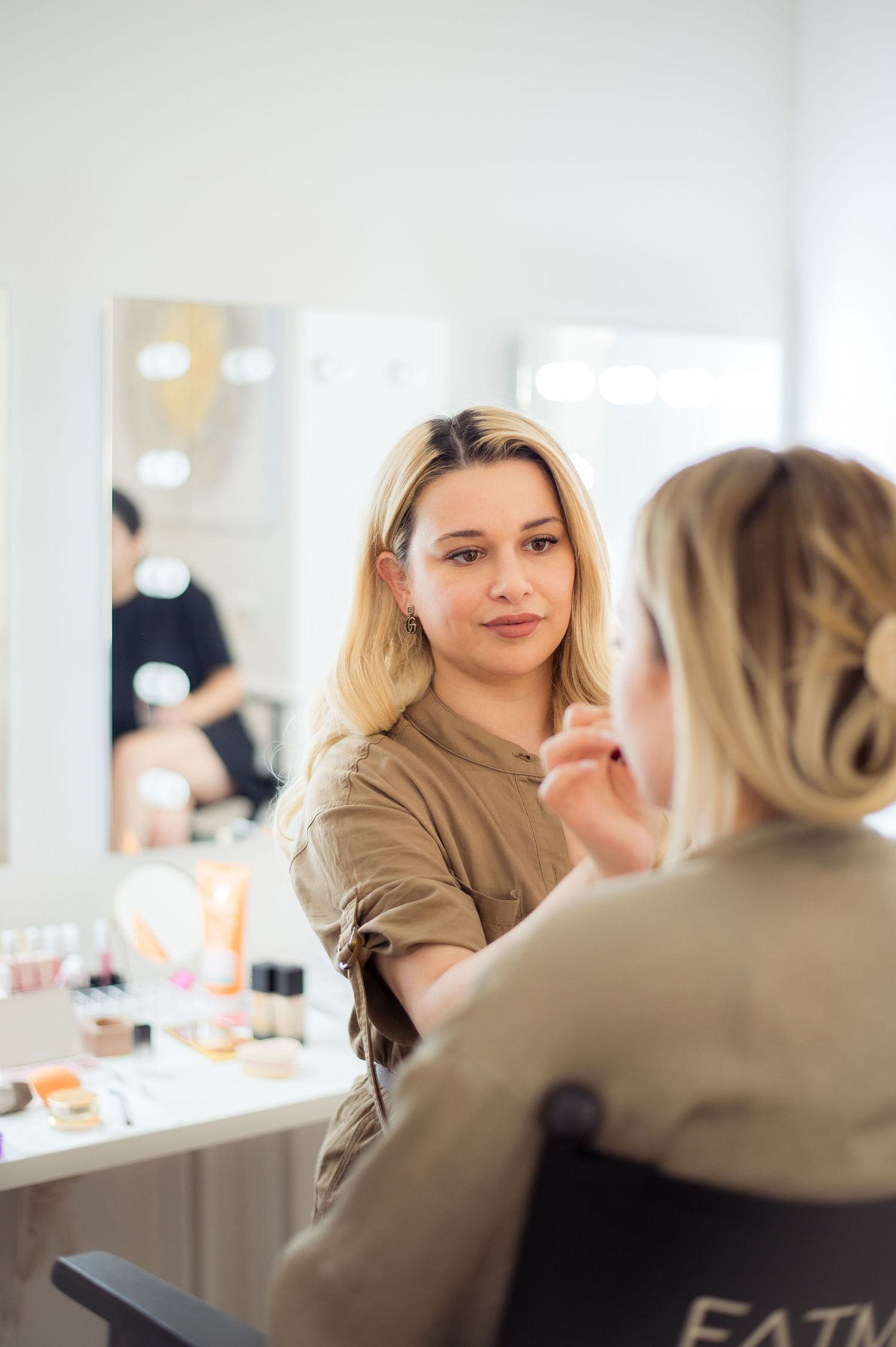 Makeup artist applying cosmetics to a blonde client in a bright vanity mirror studio setting.