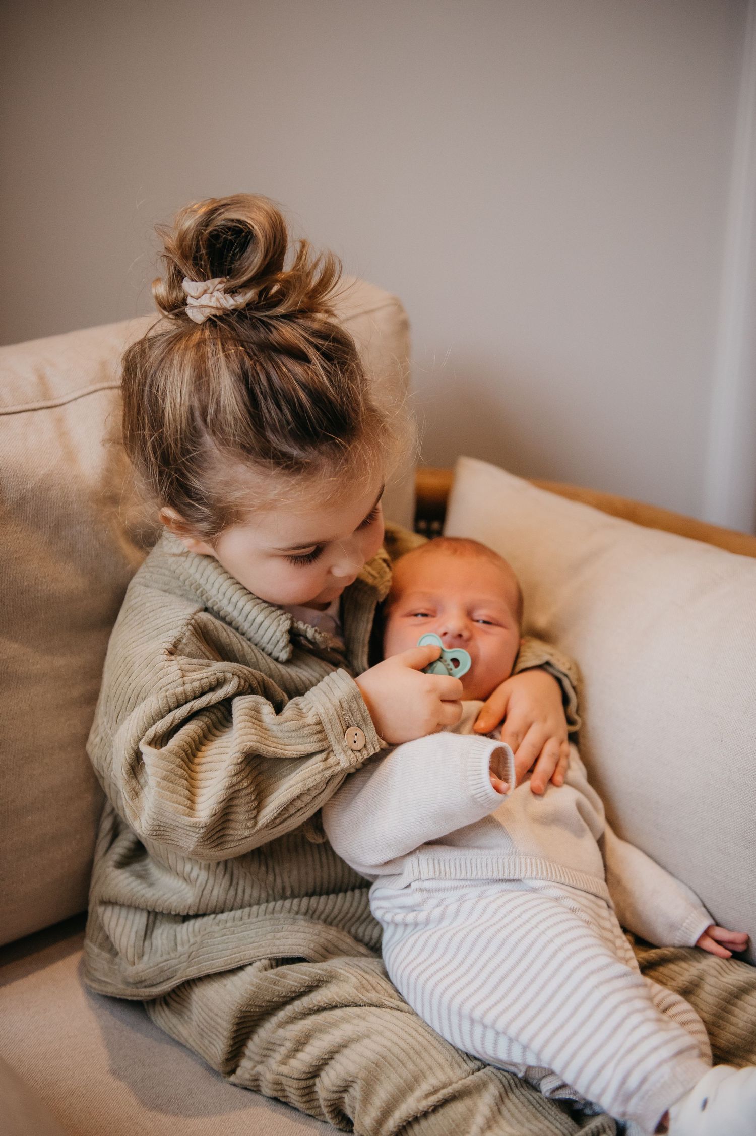 Young girl with bun hairstyle gently holds sleeping newborn baby with pacifier on a cream sofa.