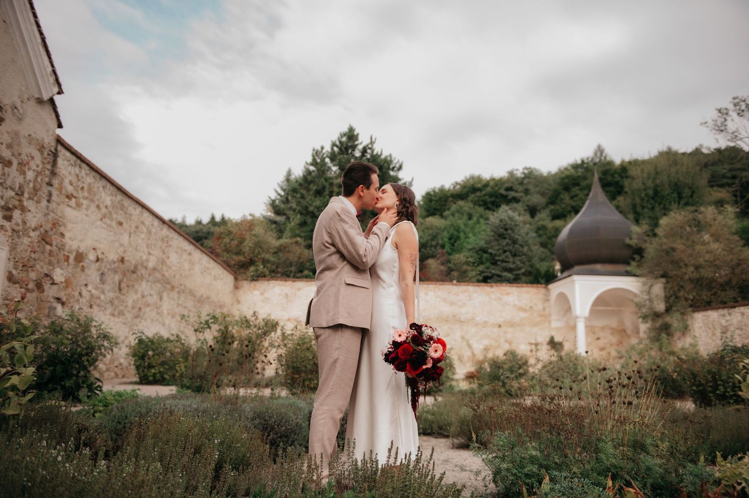 Newlywed couple sharing a kiss in a garden courtyard with stone walls and lush greenery on their wedding day.