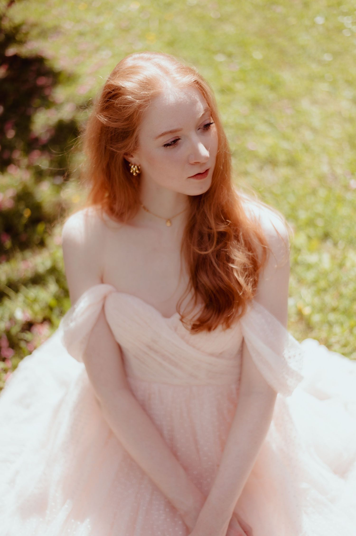 Young redhead woman in a blush pink off-shoulder tulle gown sits gracefully outdoors in sunlight.