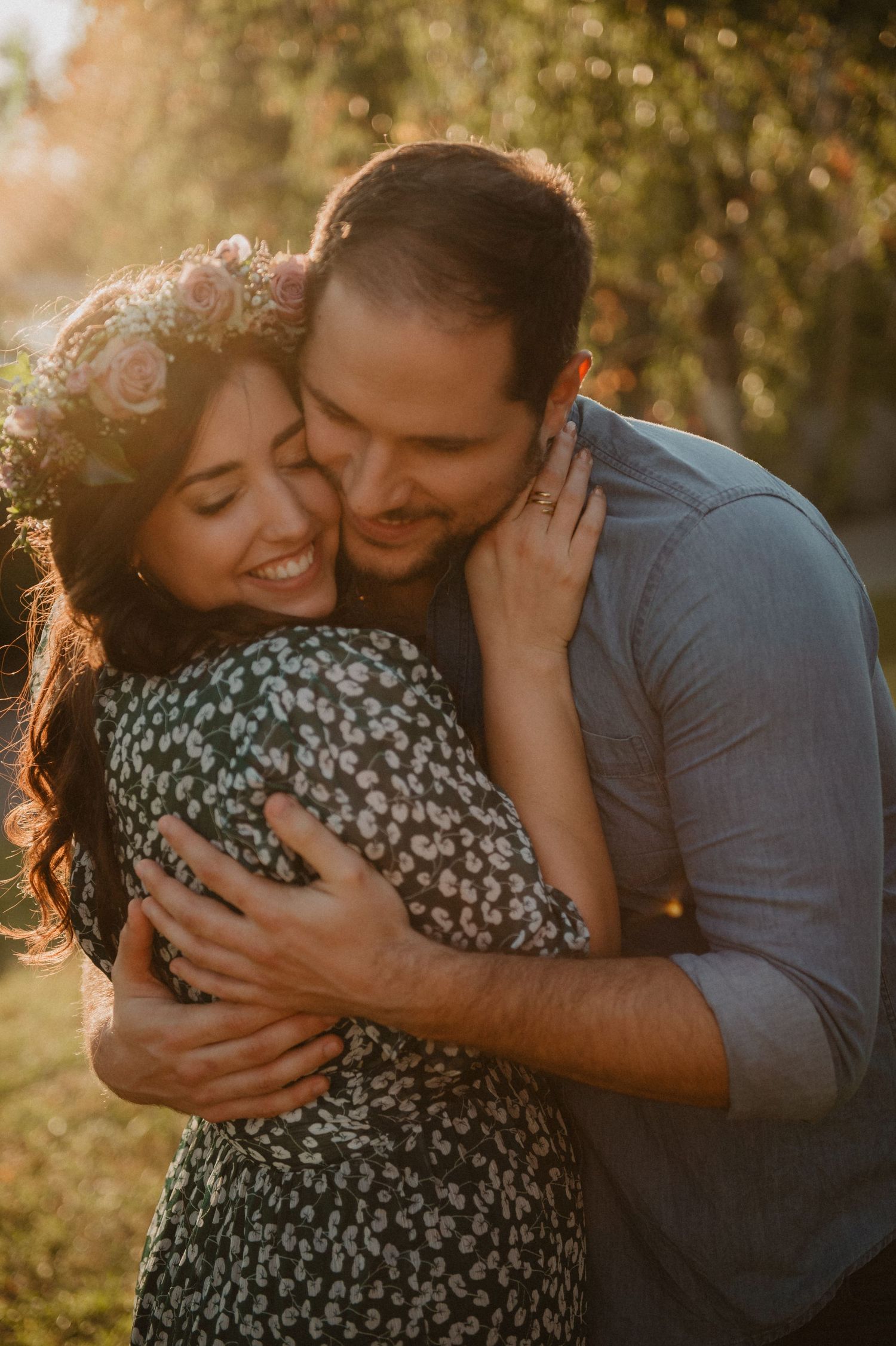 Man lovingly hugging woman from behind in warm golden sunlight, woman wearing a floral crown smiling.