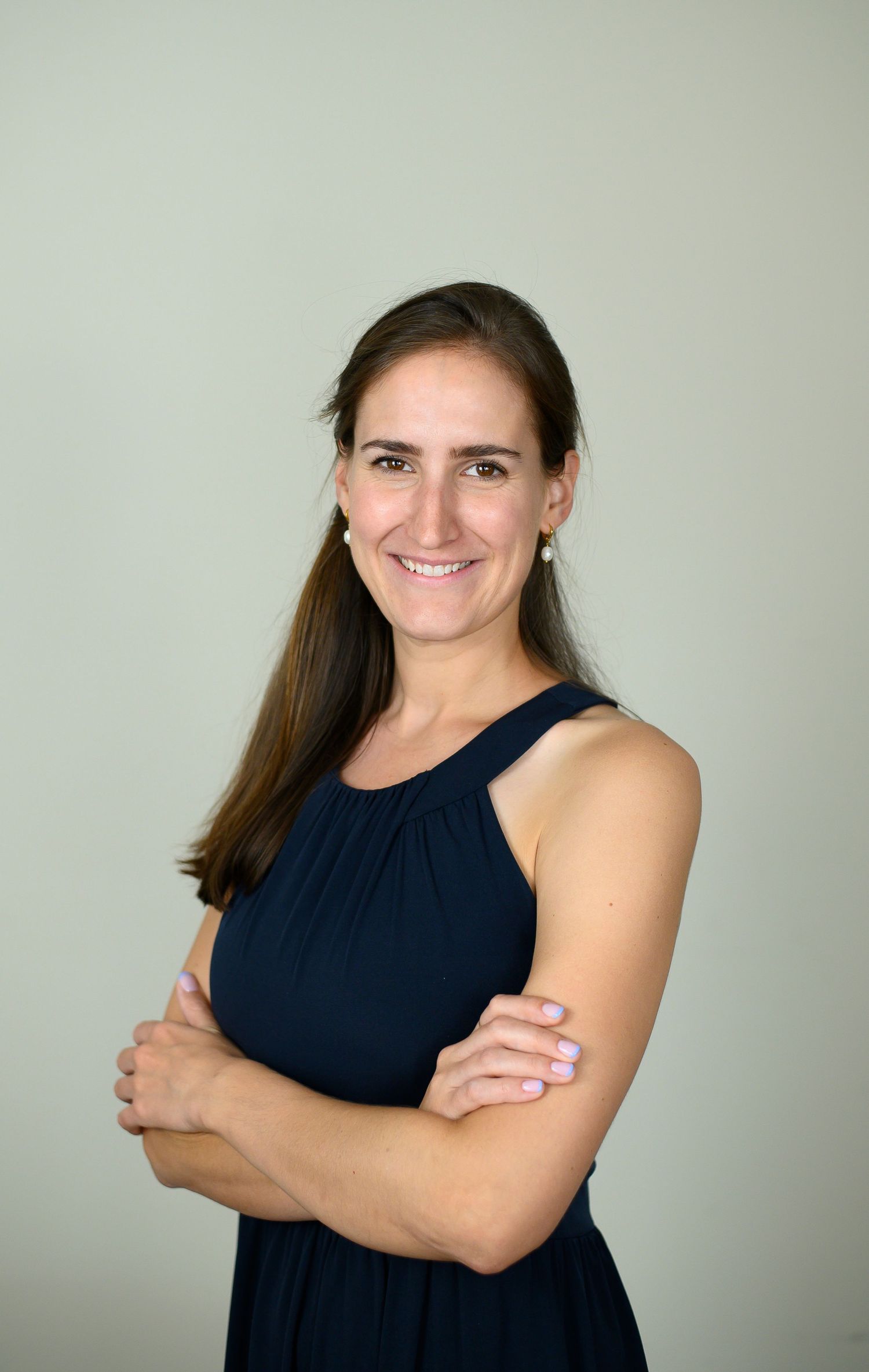 Smiling woman with long brown hair in a navy sleeveless dress, arms crossed, posing against a light gray background.