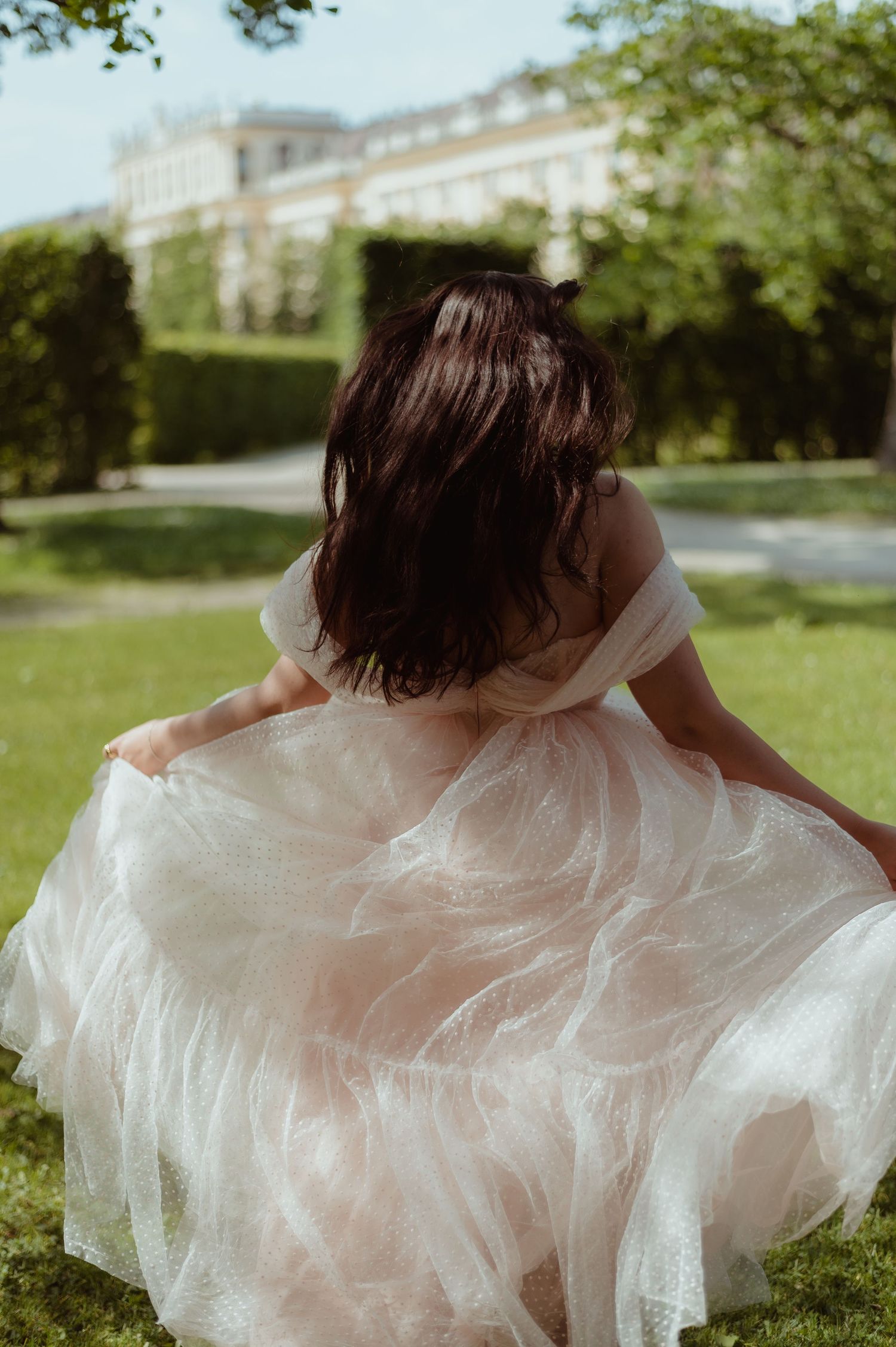 Woman in flowing white tulle gown twirling in a lush garden with a grand building in the background.