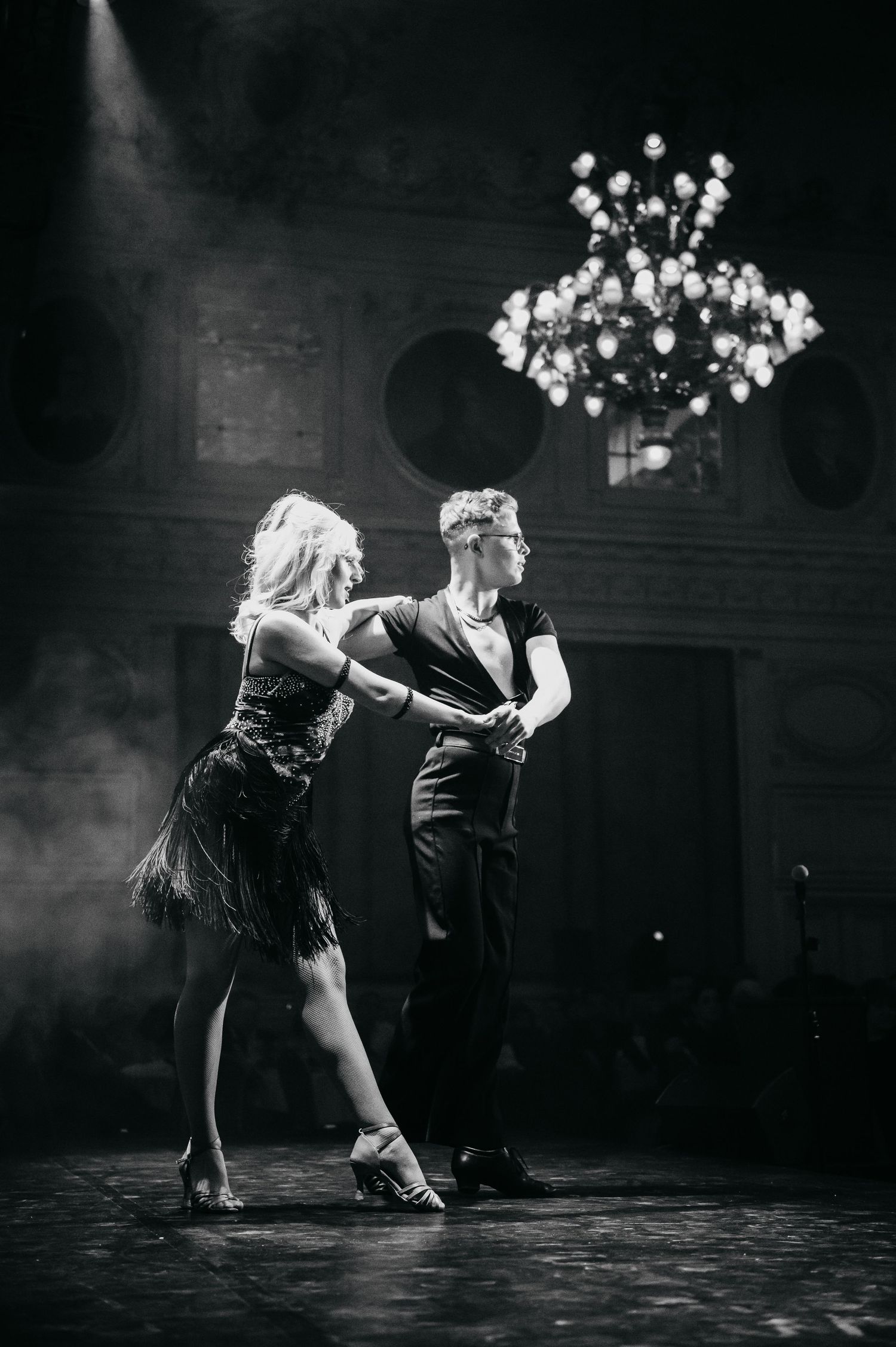 Elegant couple dancing ballroom in black and white under chandelier with dramatic stage lighting and smoke.