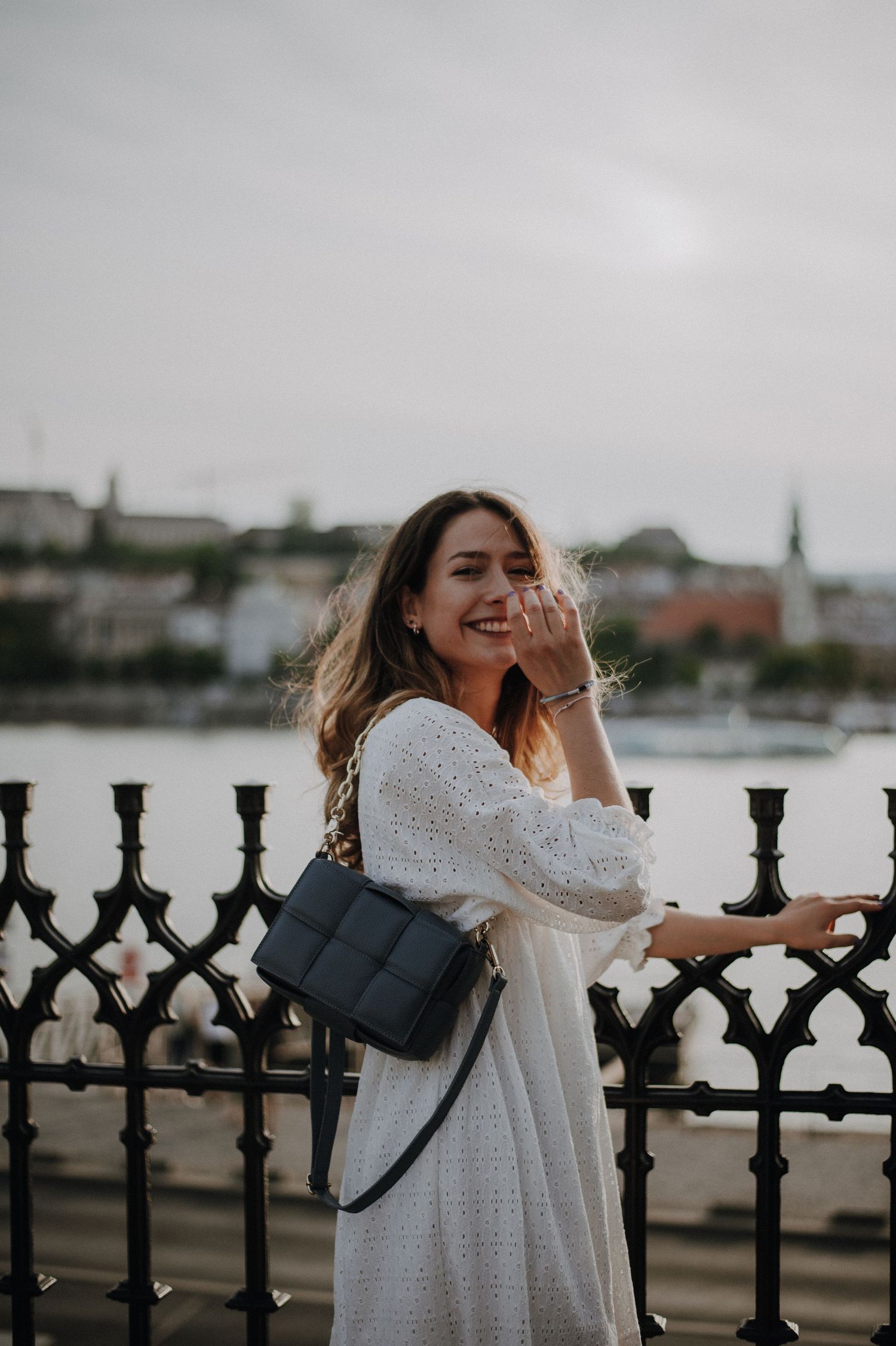 Smiling woman in white dress with black quilted bag posing by ornate iron railing near waterfront.