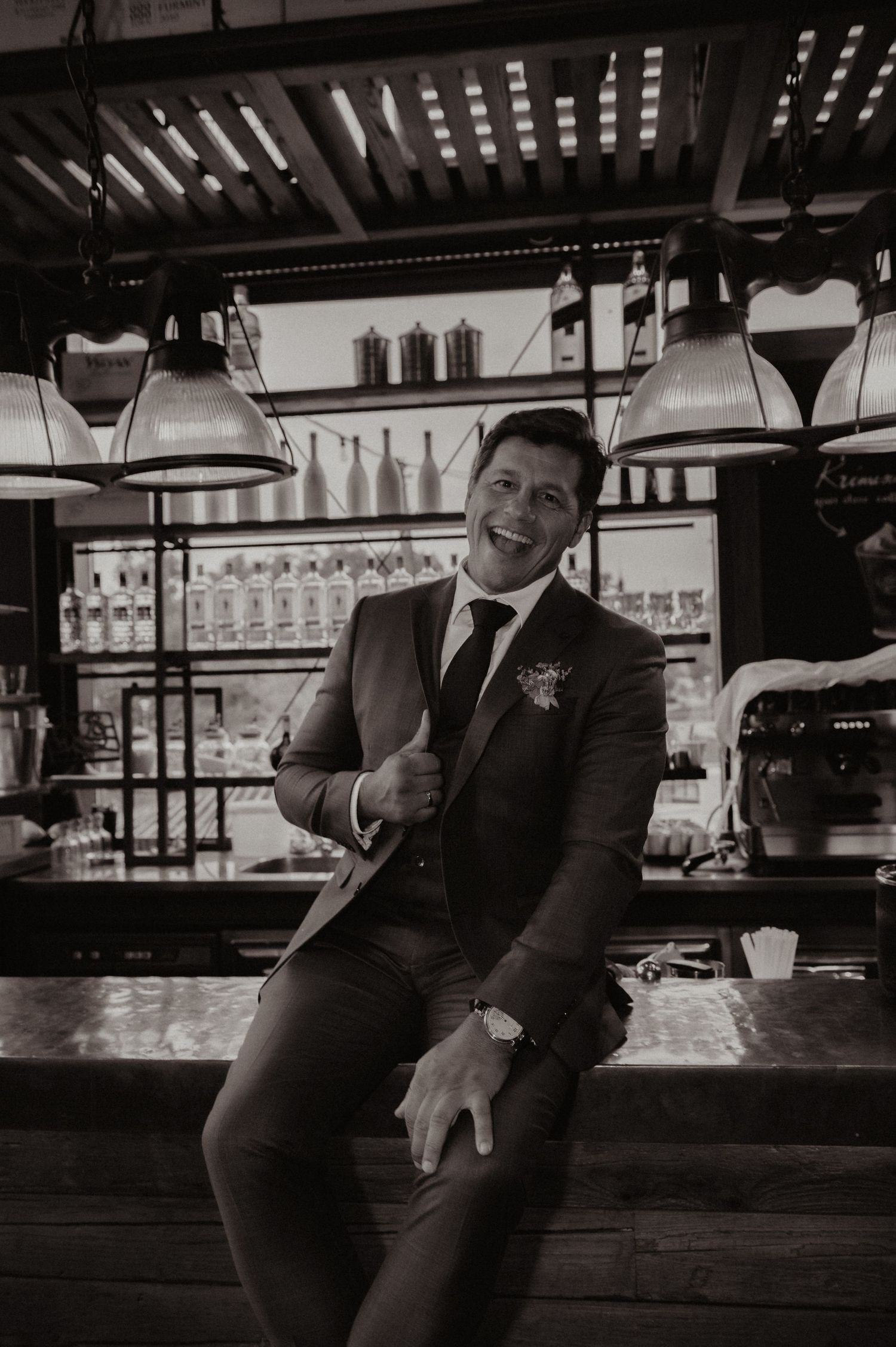 Smiling groom in dark suit with boutonniere sits at bar in black and white wedding photo.