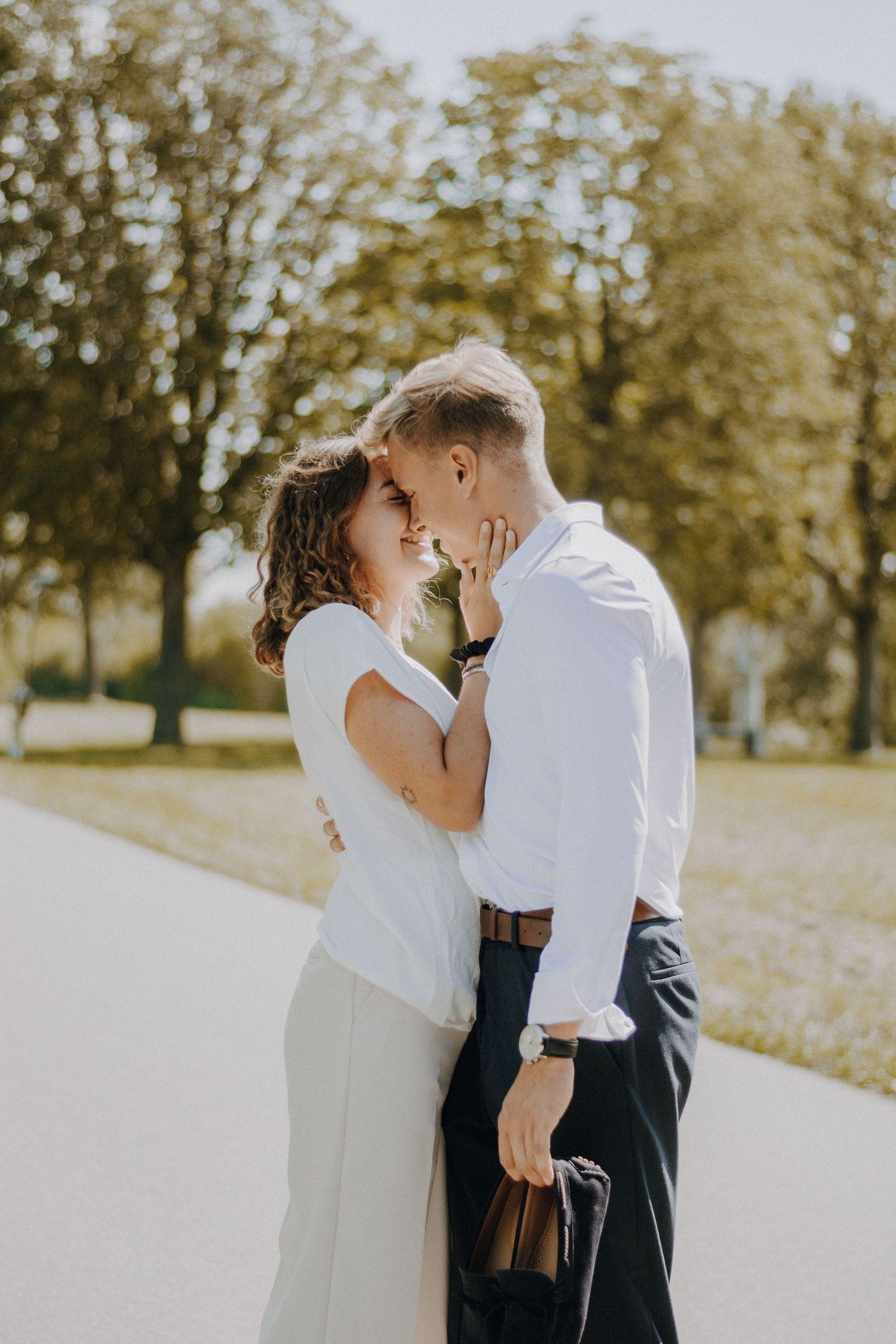 Couple sharing a romantic moment outdoors, leaning in for a kiss in a sunny park with lush green trees in the background.