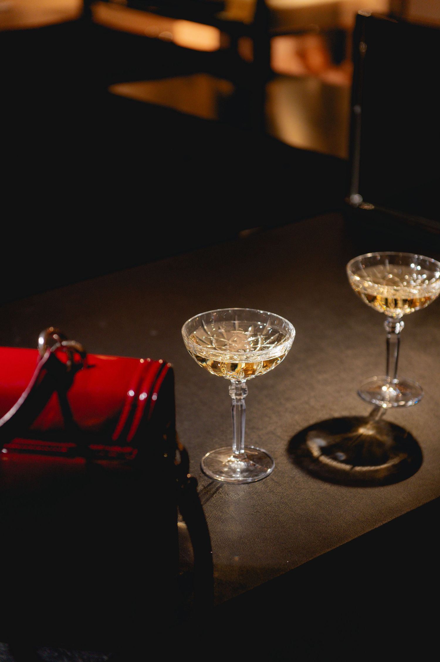 Two elegant crystal coupe glasses with champagne on a dark table beside a red folded napkin in a dimly lit bar.