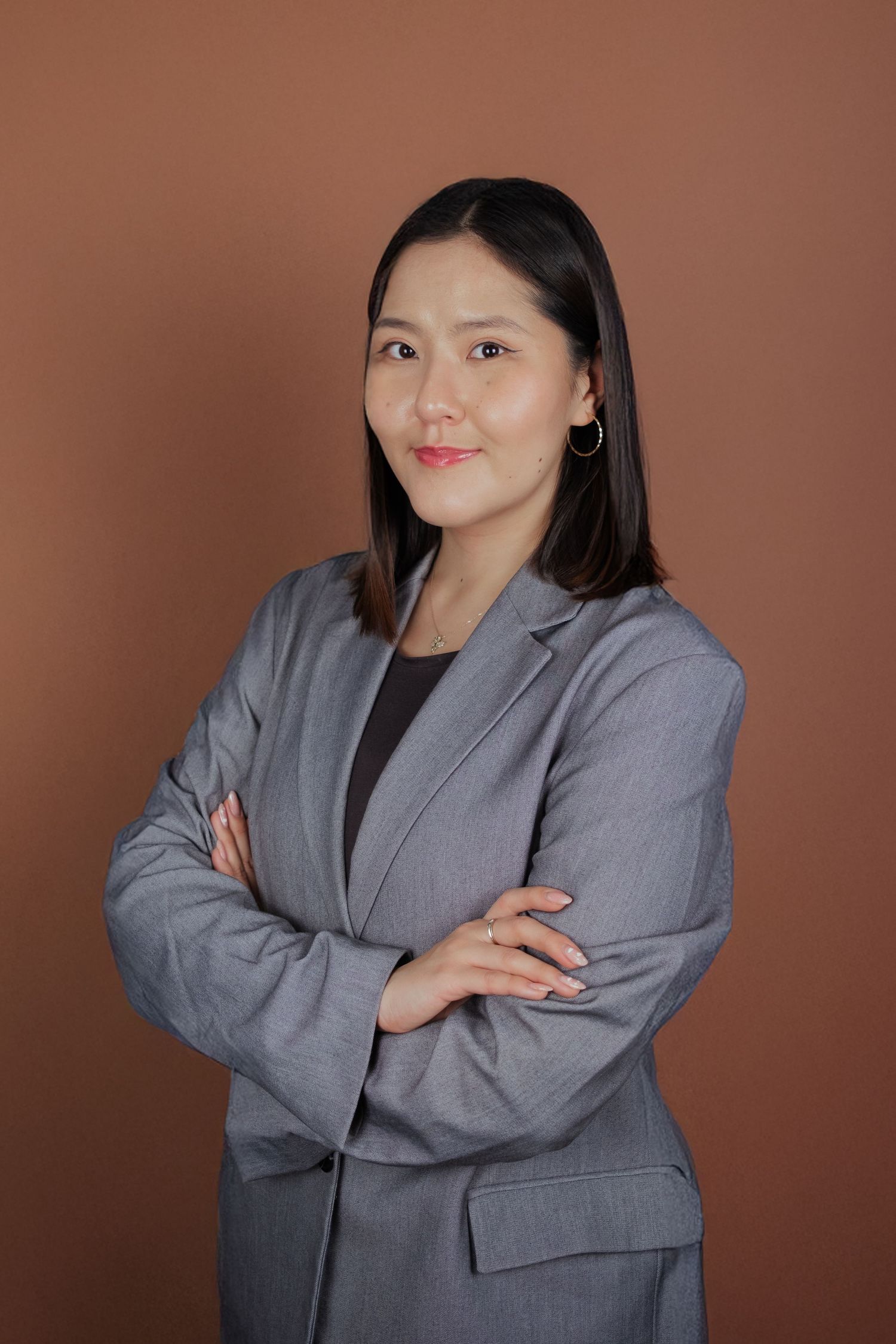 Professional Asian businesswoman in gray blazer with arms crossed, smiling confidently against brown background.