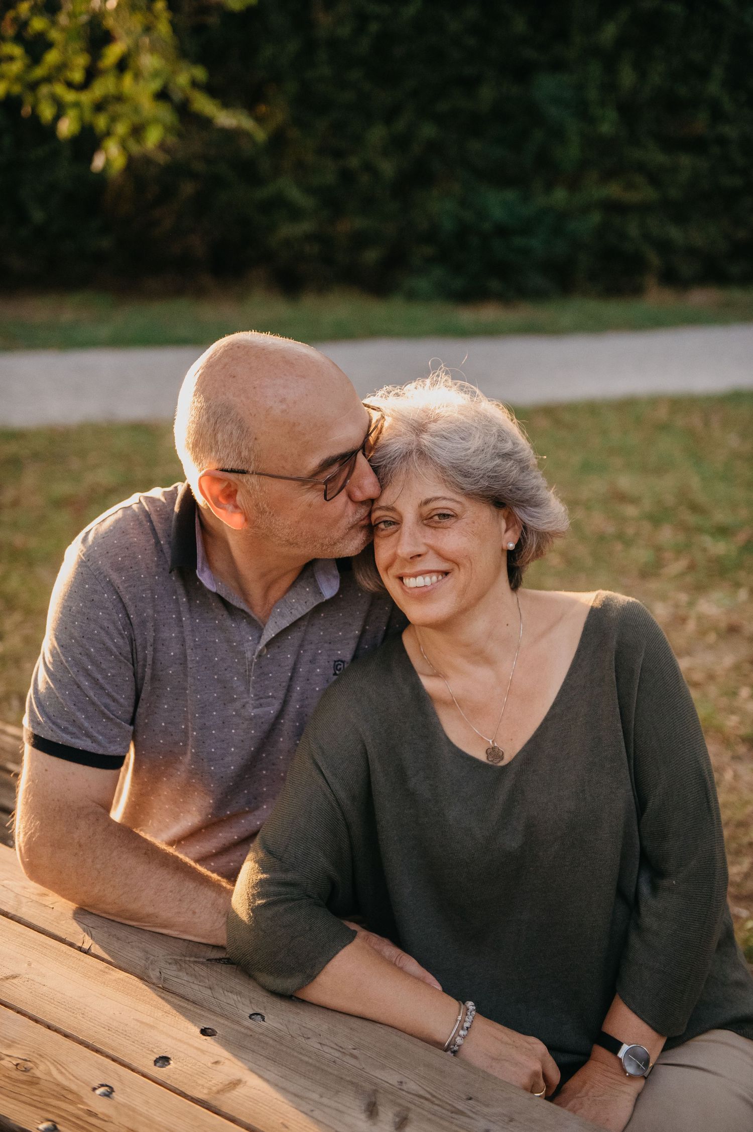 An older couple sitting at a picnic table outdoors, man kissing woman's cheek as she smiles warmly.