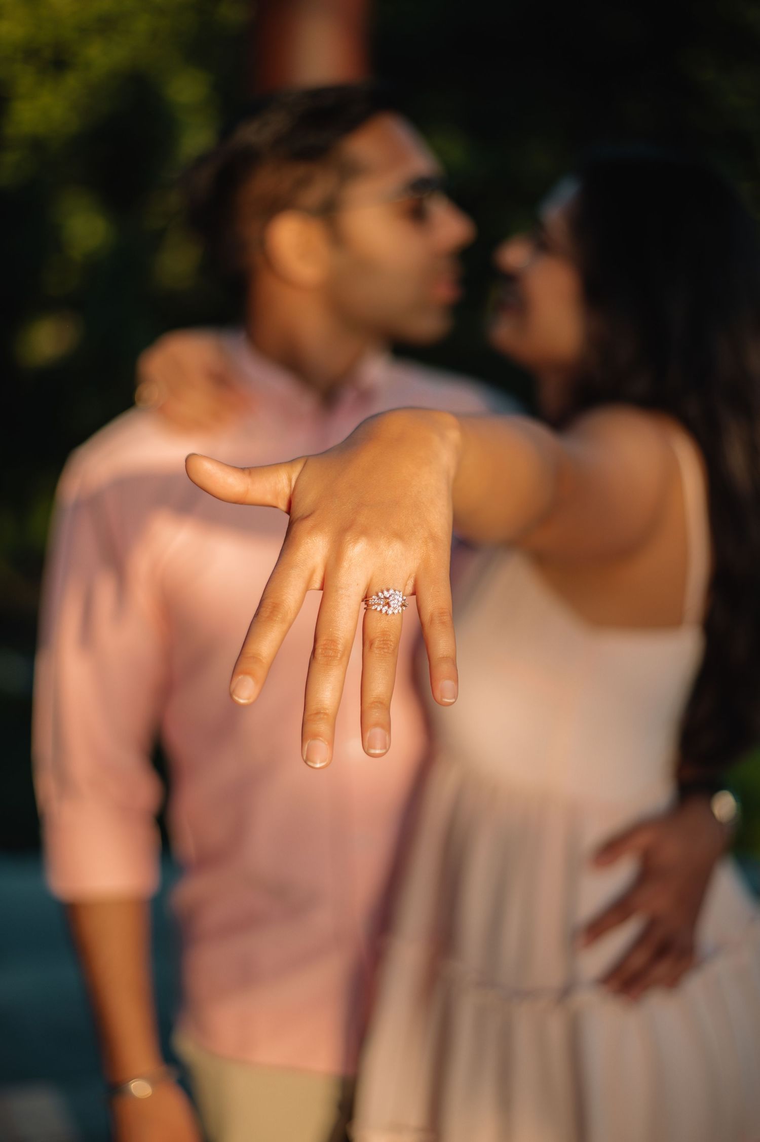 Couple sharing a romantic moment while woman displays sparkling engagement ring toward camera in soft evening light.