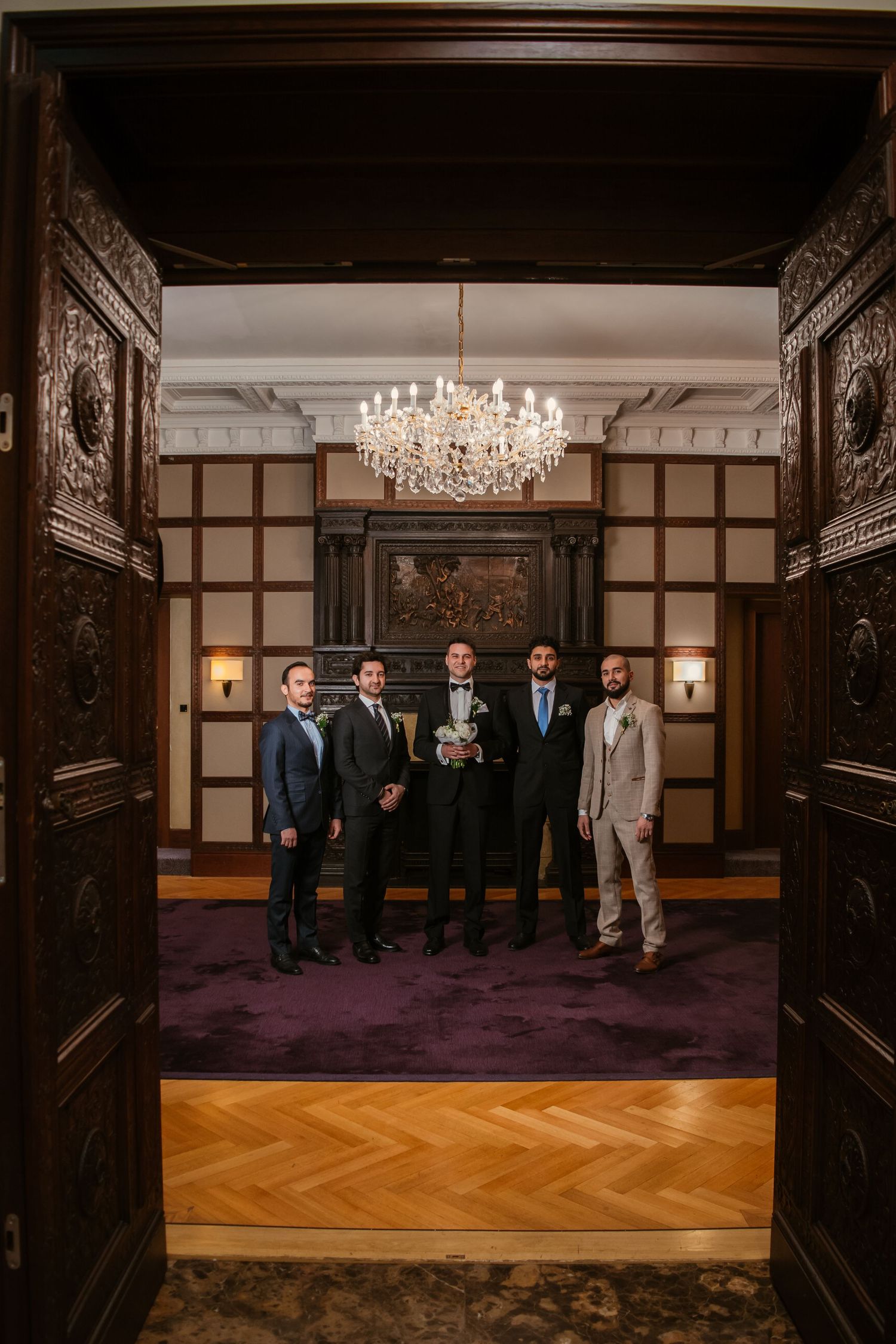 Groom and groomsmen in suits pose under crystal chandelier in elegant wood-paneled room with purple carpet.