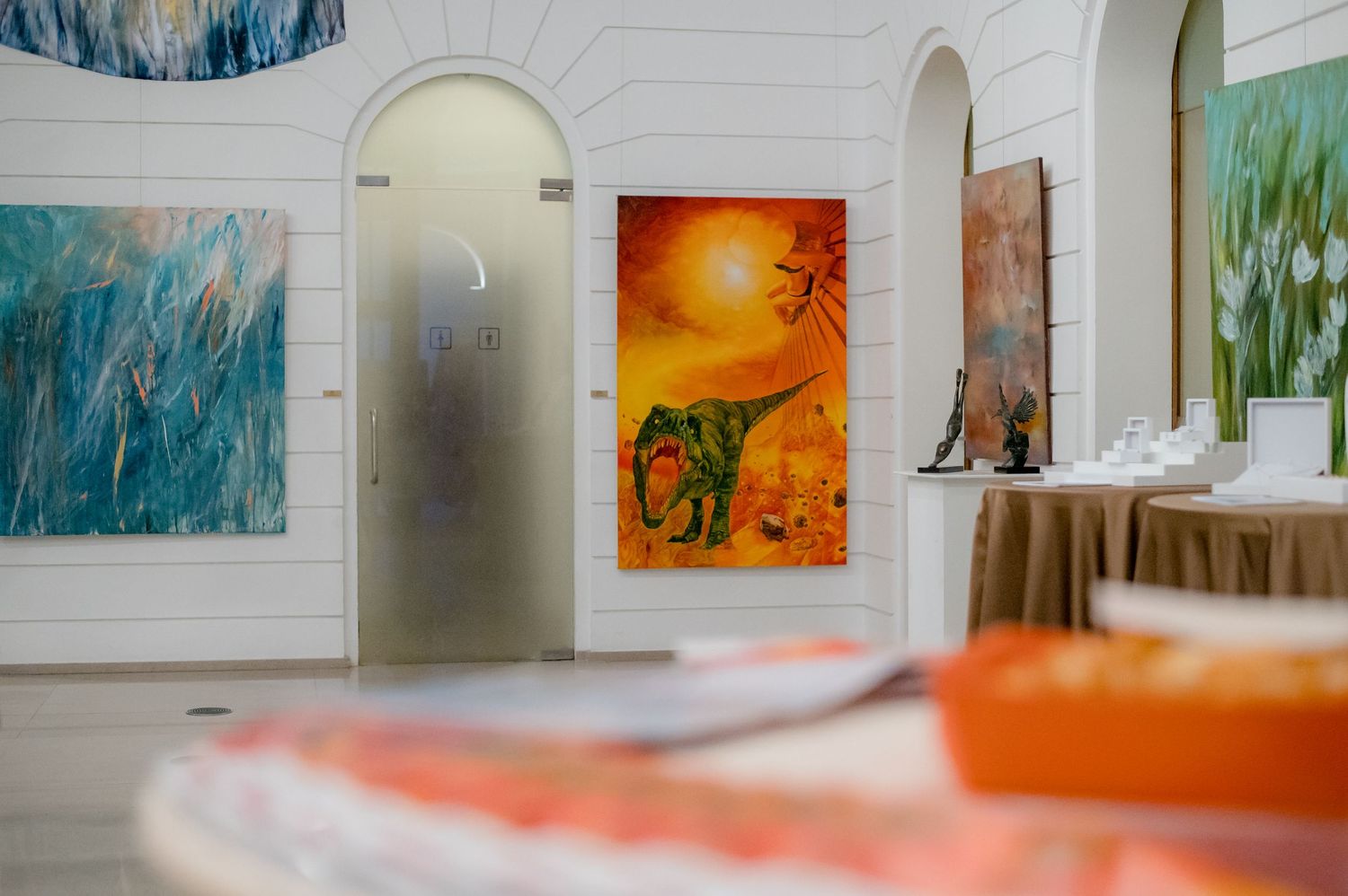 Art gallery interior with vibrant paintings on white walls, featuring orange landscape and blue abstract artworks.