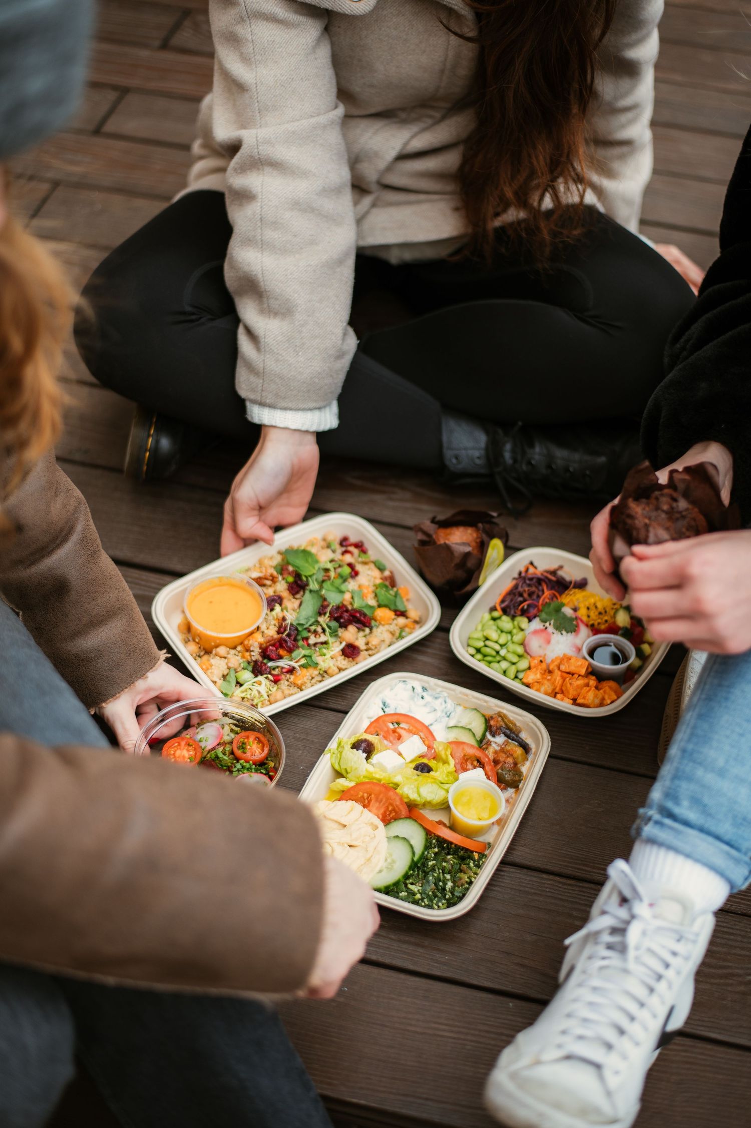 Group of friends sitting on wooden deck sharing colorful healthy salad bowls with fresh vegetables outdoors.
