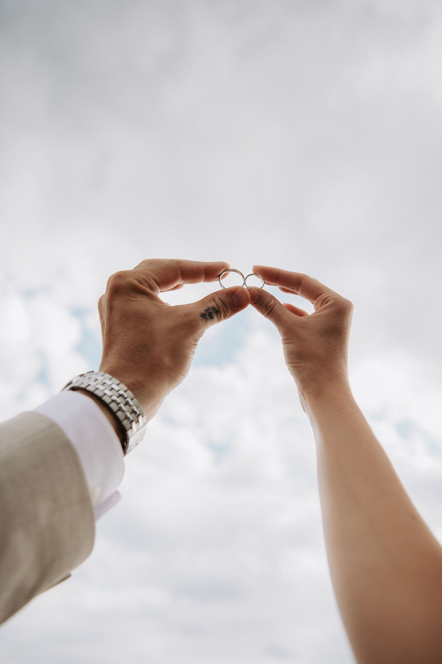 Couple holding two gold wedding rings together against a cloudy sky backdrop.