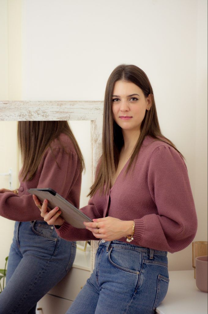 Brunette woman in a mauve knit sweater and jeans holds a tablet near a white mirror indoors.