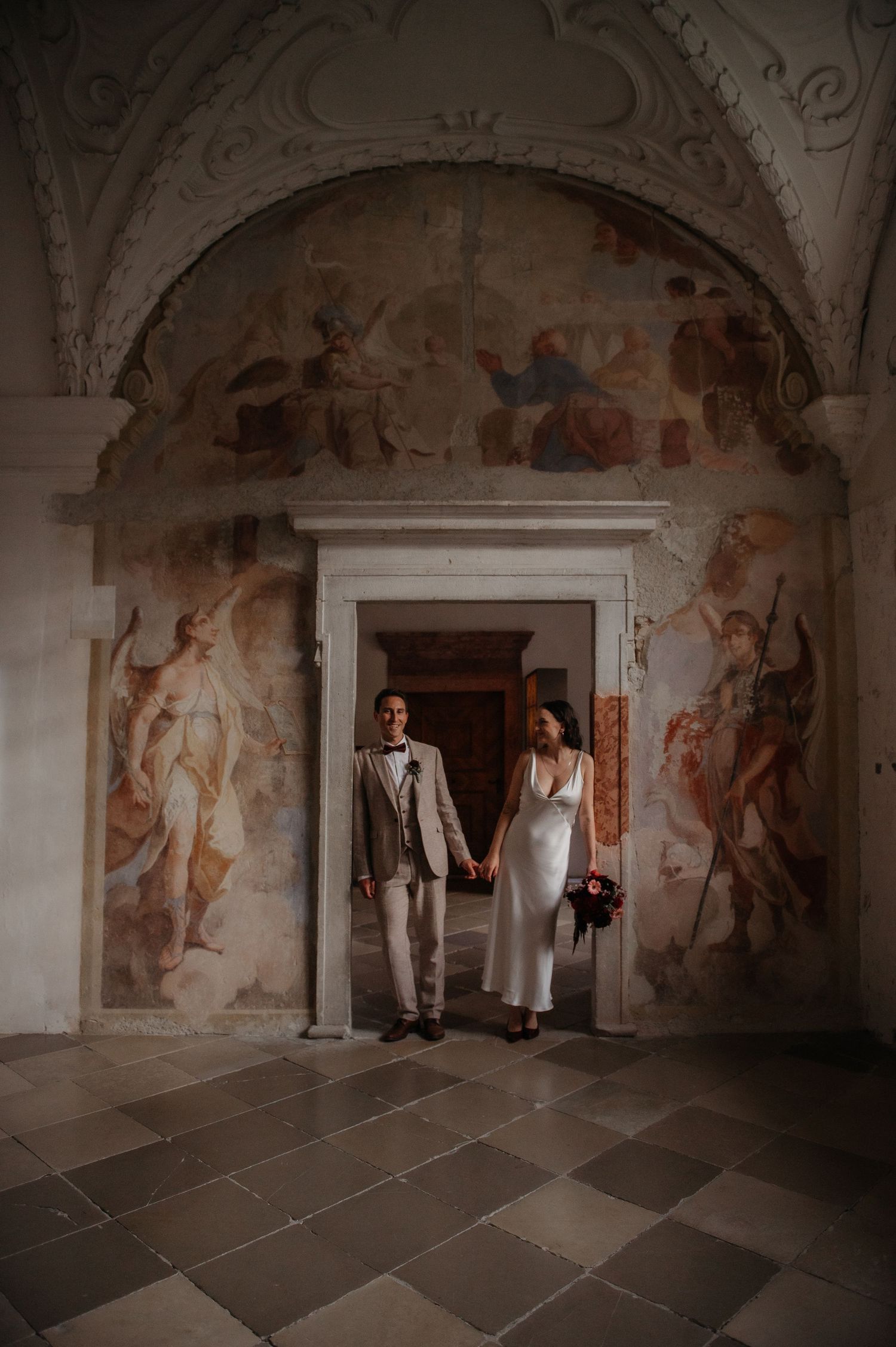 Bride and groom hold hands in elegant venue with ornate Renaissance frescoes and arched doorway on their wedding day.