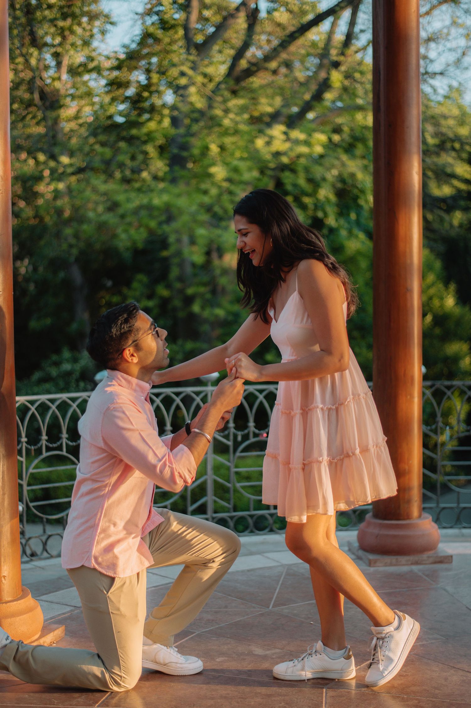 Man kneeling on one knee proposing to a woman in a pink dress at a scenic outdoor gazebo surrounded by lush greenery.