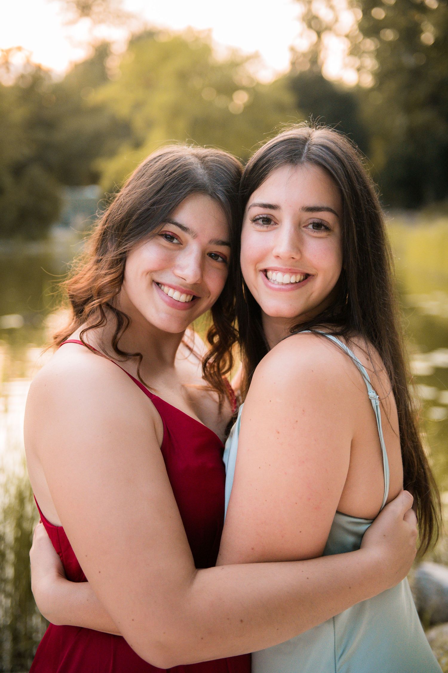 Two young women in formal dresses hugging and smiling outdoors near a pond with golden hour lighting.