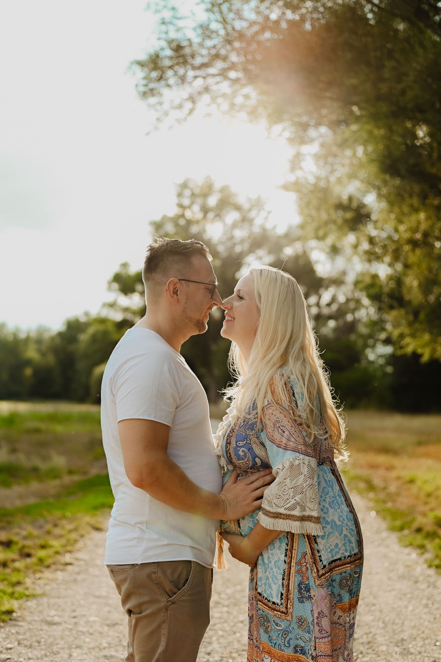 A romantic couple shares an intimate moment on a sunlit path surrounded by lush green trees during golden hour.