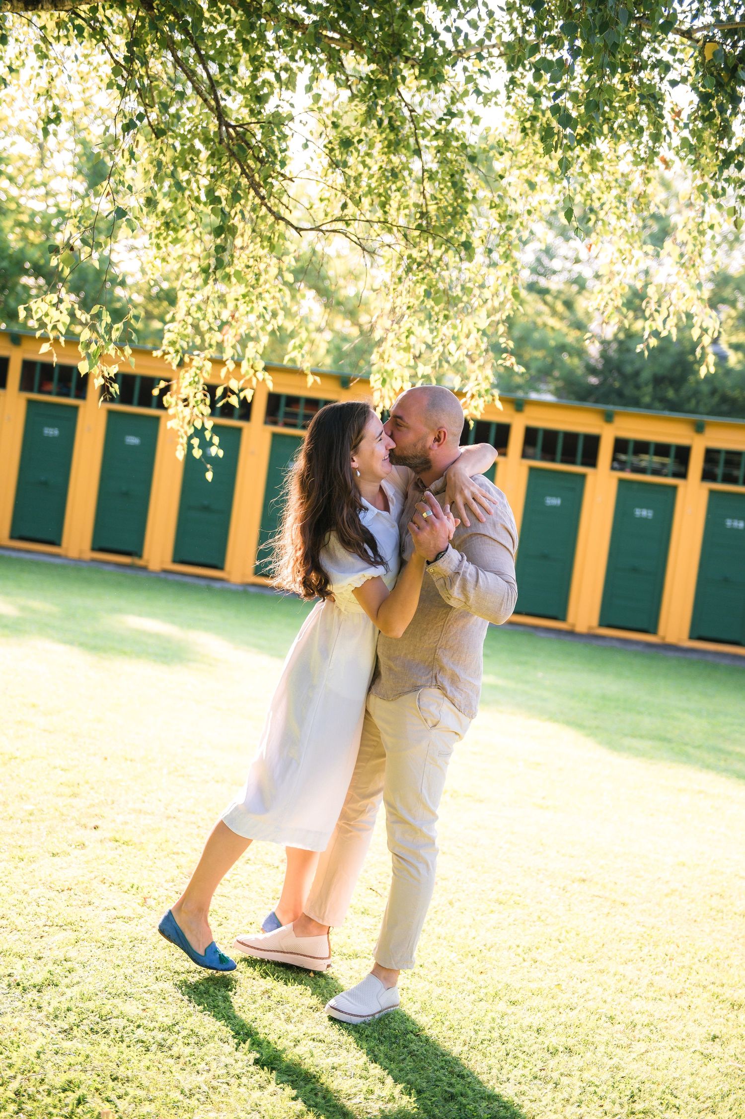 Couple sharing a romantic kiss under a tree on a sunny day with yellow and green cabanas in the background.
