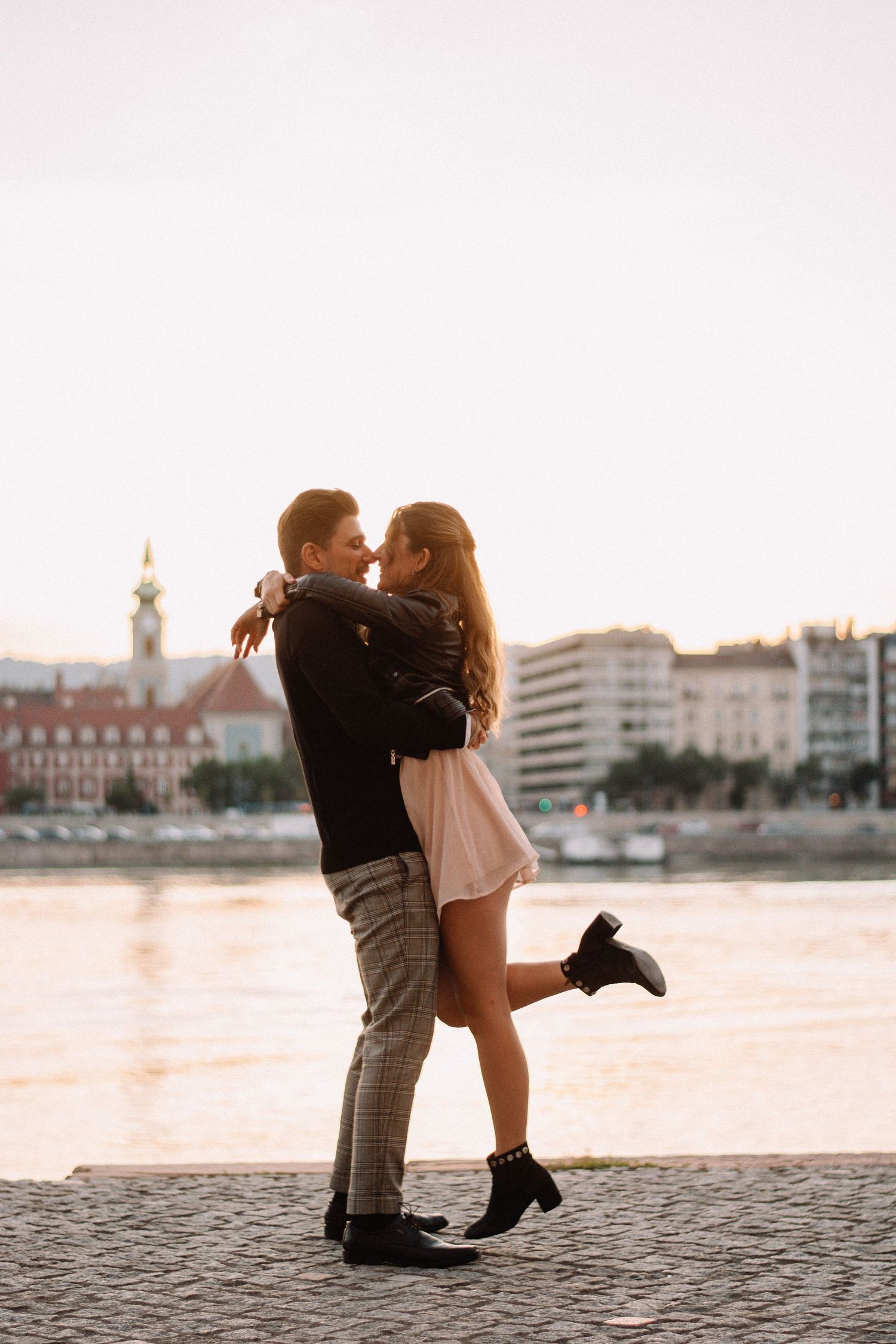 Romantic couple embracing near a riverside at sunset with historic buildings in the background.