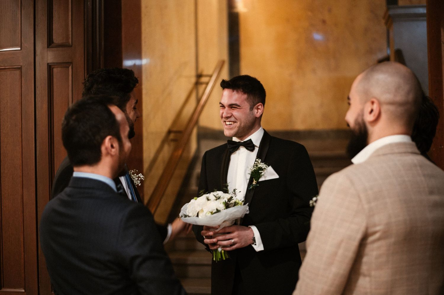 Smiling groom in black tuxedo holding white bouquet with groomsmen in ornate wedding venue.